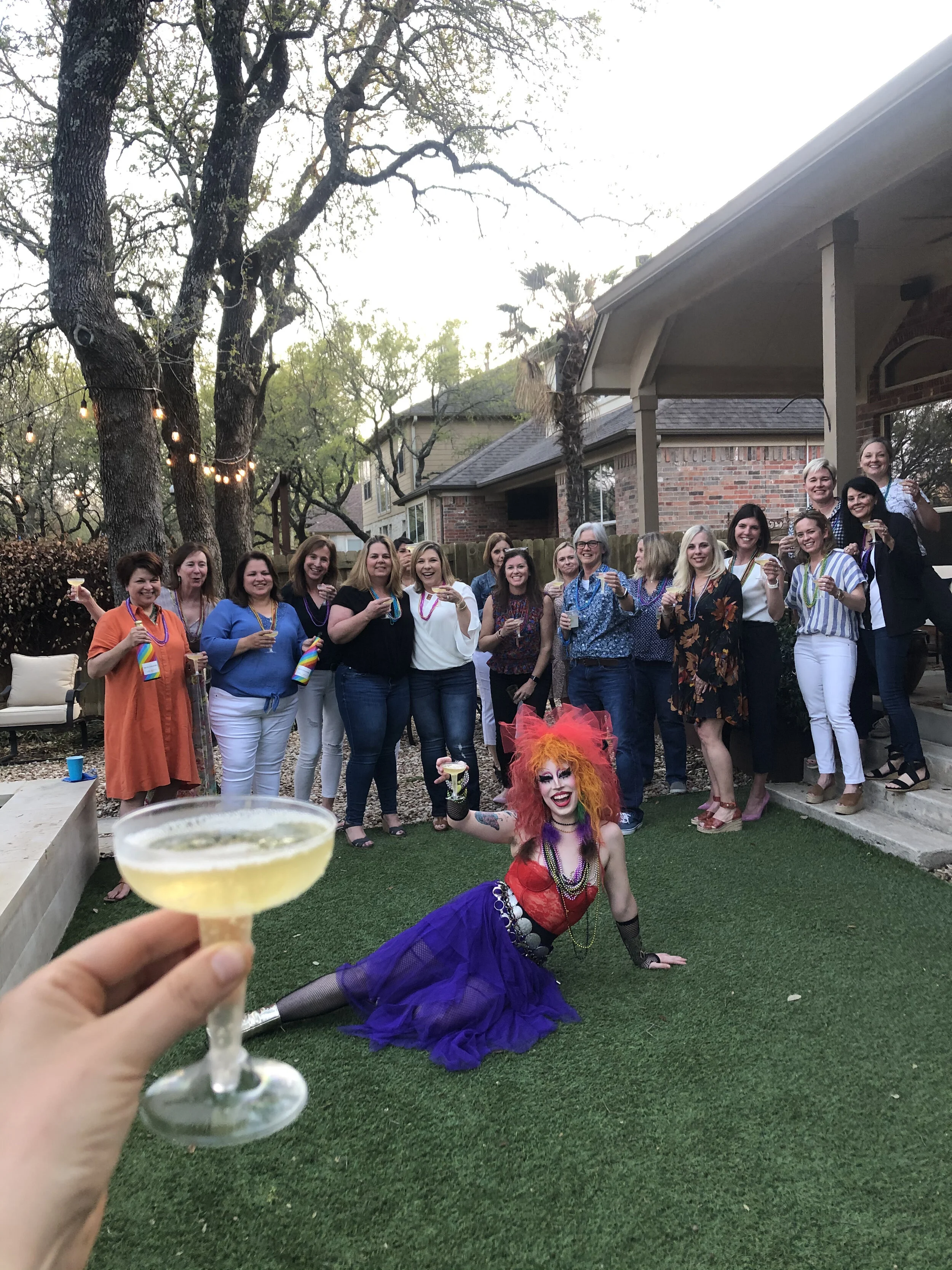 A group of women celebrating outdoors at a backyard party, with a performer dressed as a colorful clown or carnival performer sitting on the grass in front. The women are smiling and holding drinks, and one person is holding a cocktail glass in the f