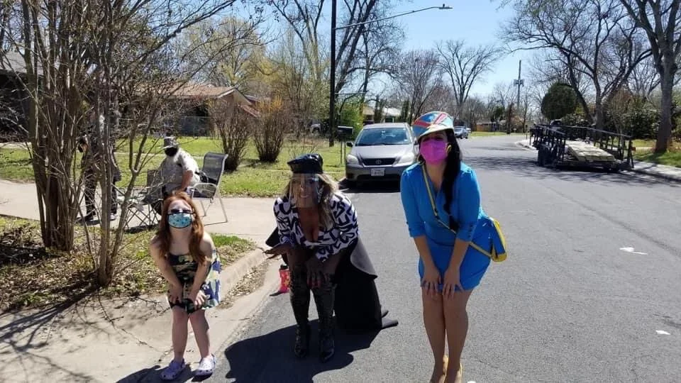 Four people wearing masks and hats standing on a street, with trees, grass, parked cars, and a street setup in the background.