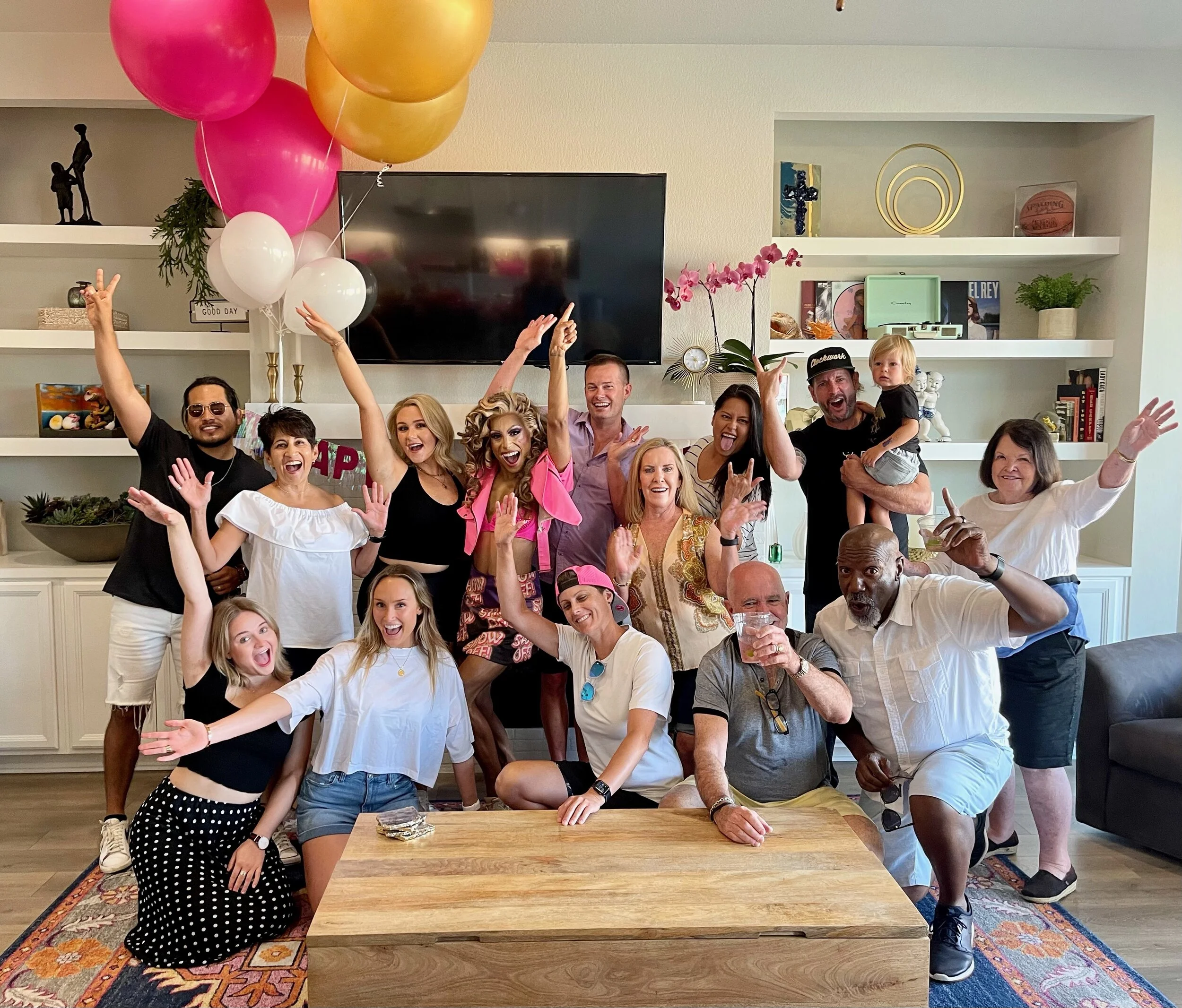 A group of people celebrating indoors, with some holding balloons, making peace signs, and smiling. There is a mix of men, women, and children, suggesting a festive gathering or party.