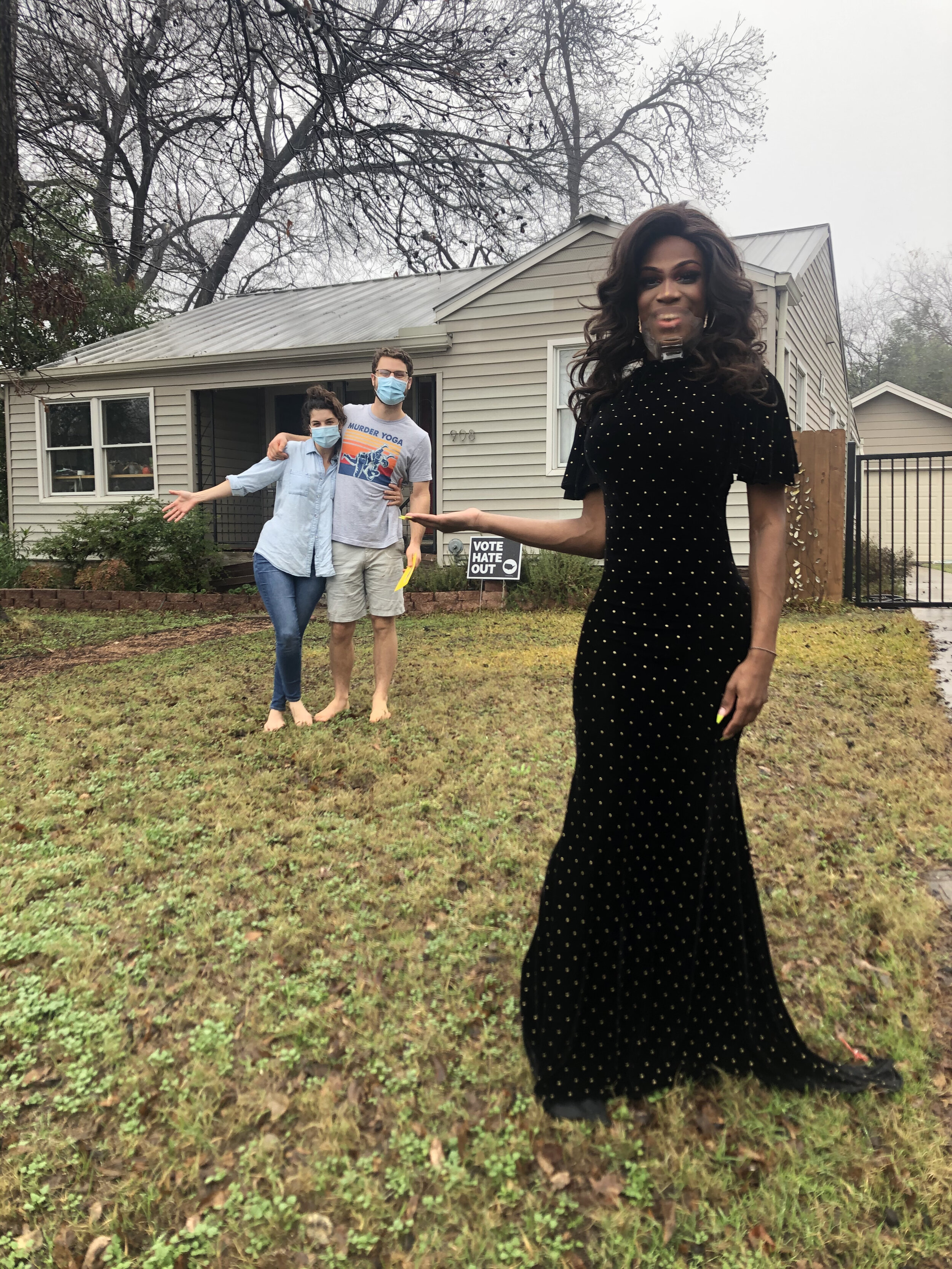 A person in a long black dress with yellow polka dots stands on the grass in front of a house, with two people in masks in the background near the house, and a yard sign that reads 'Vote Hate Out'.