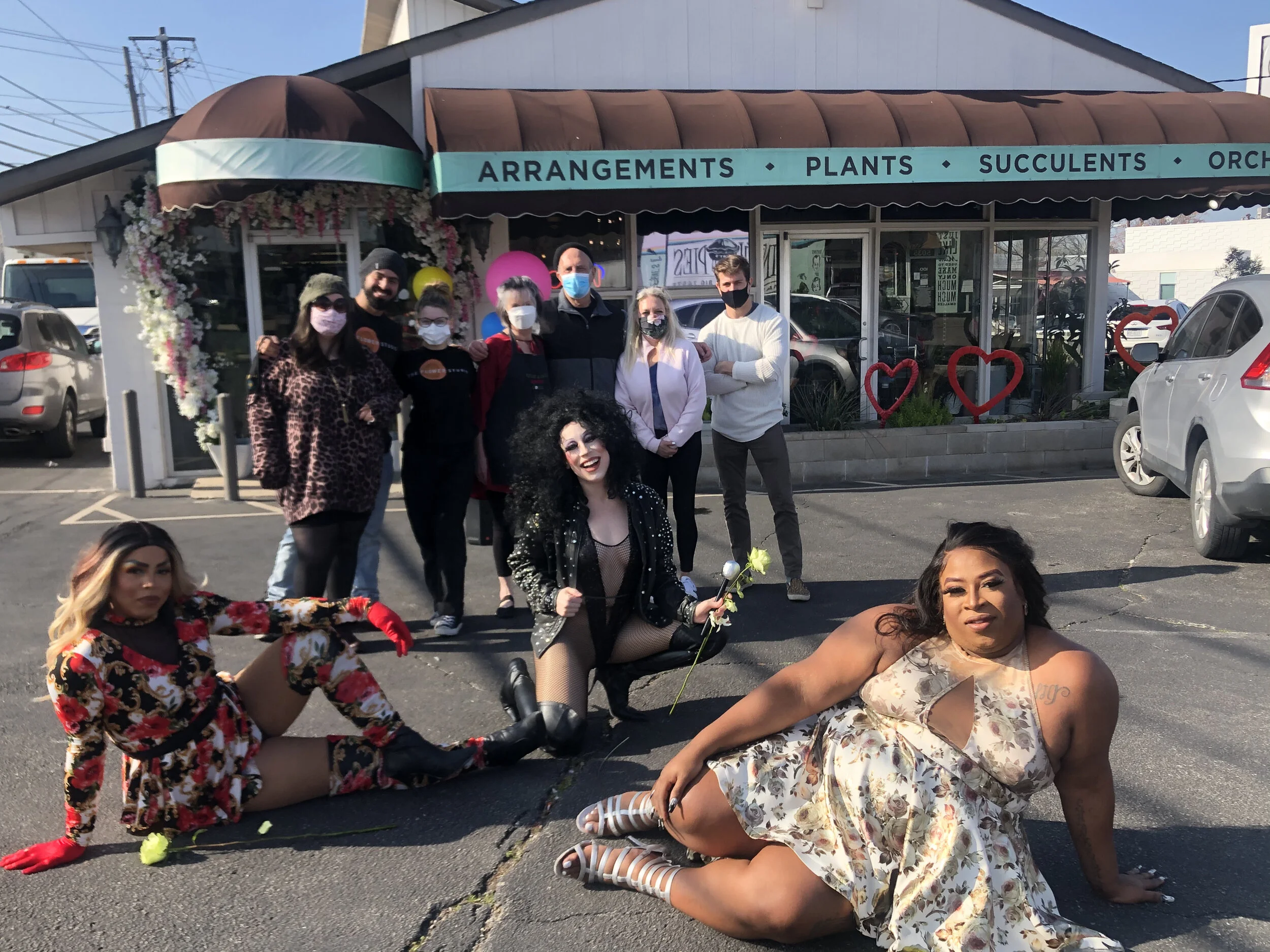 Group of nine people, including three women in the front sitting or kneeling on the ground and six people standing behind them, posing for a photo outside a flower shop decorated with hearts and balloons. The women in the front wear colorful outfits,