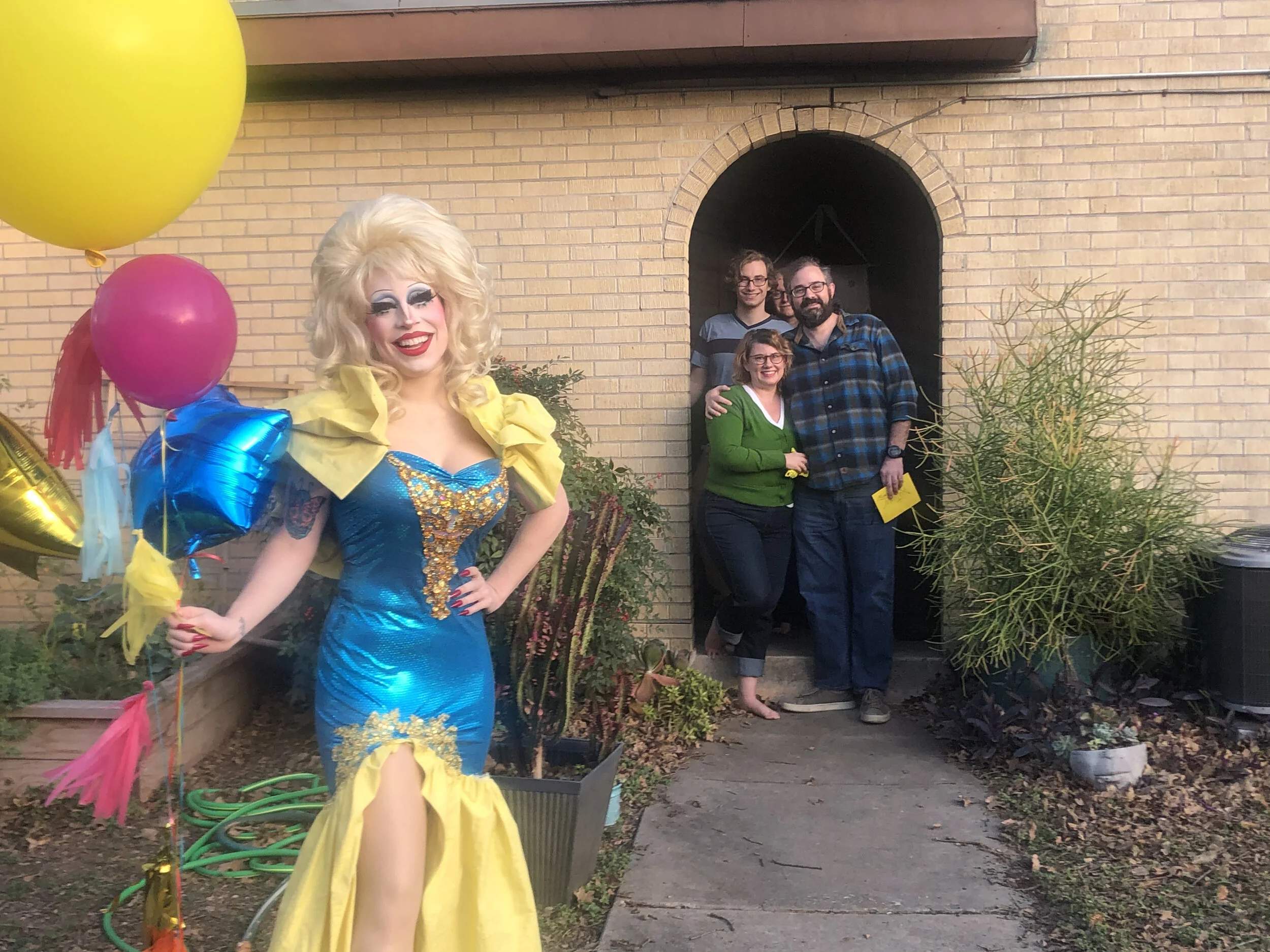 A person dressed as a drag queen in a blue and yellow dress with platinum blonde hair, holding balloons, standing outside near a group of three smiling people in casual clothing at their home.