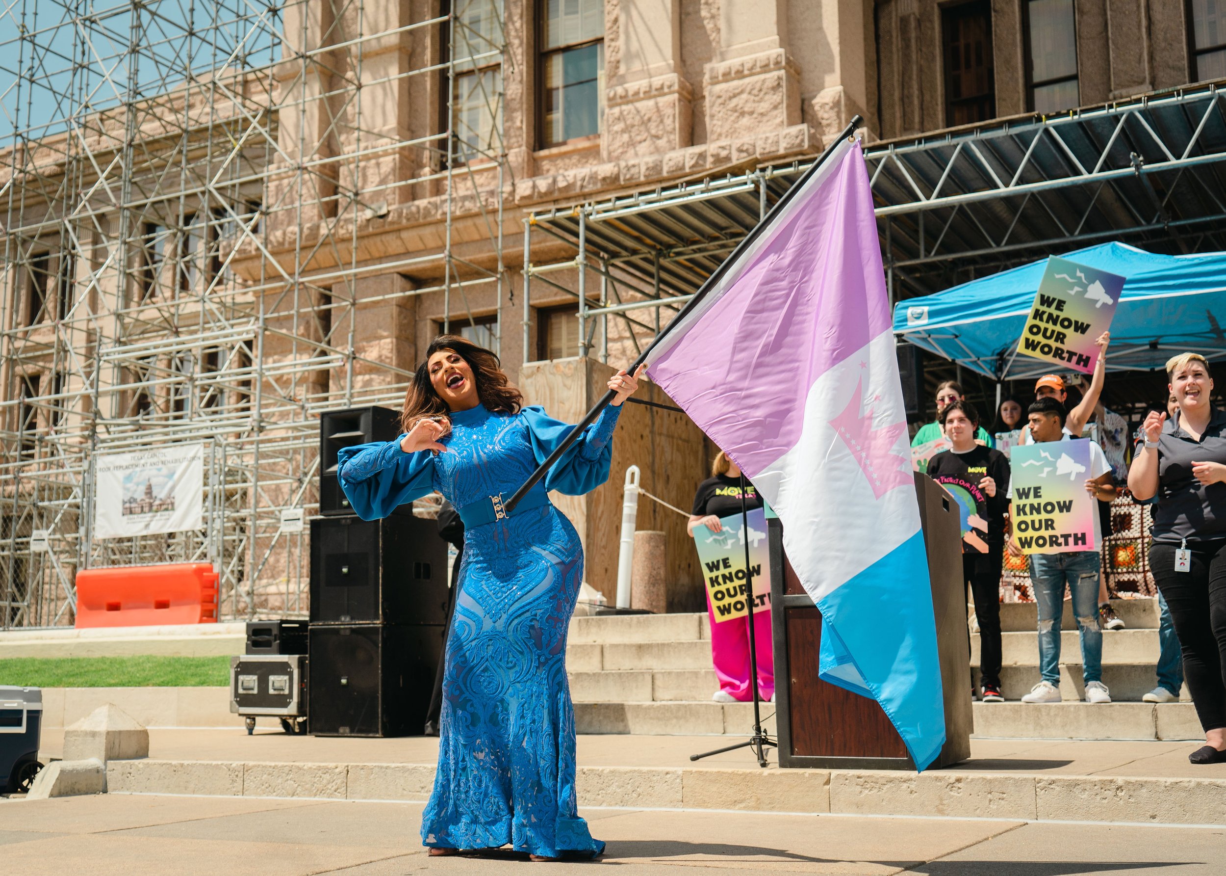 A woman in a blue dress waving a flag at a rally or protest with people holding signs that say "We know our worth" in front of a historic building with scaffolding.