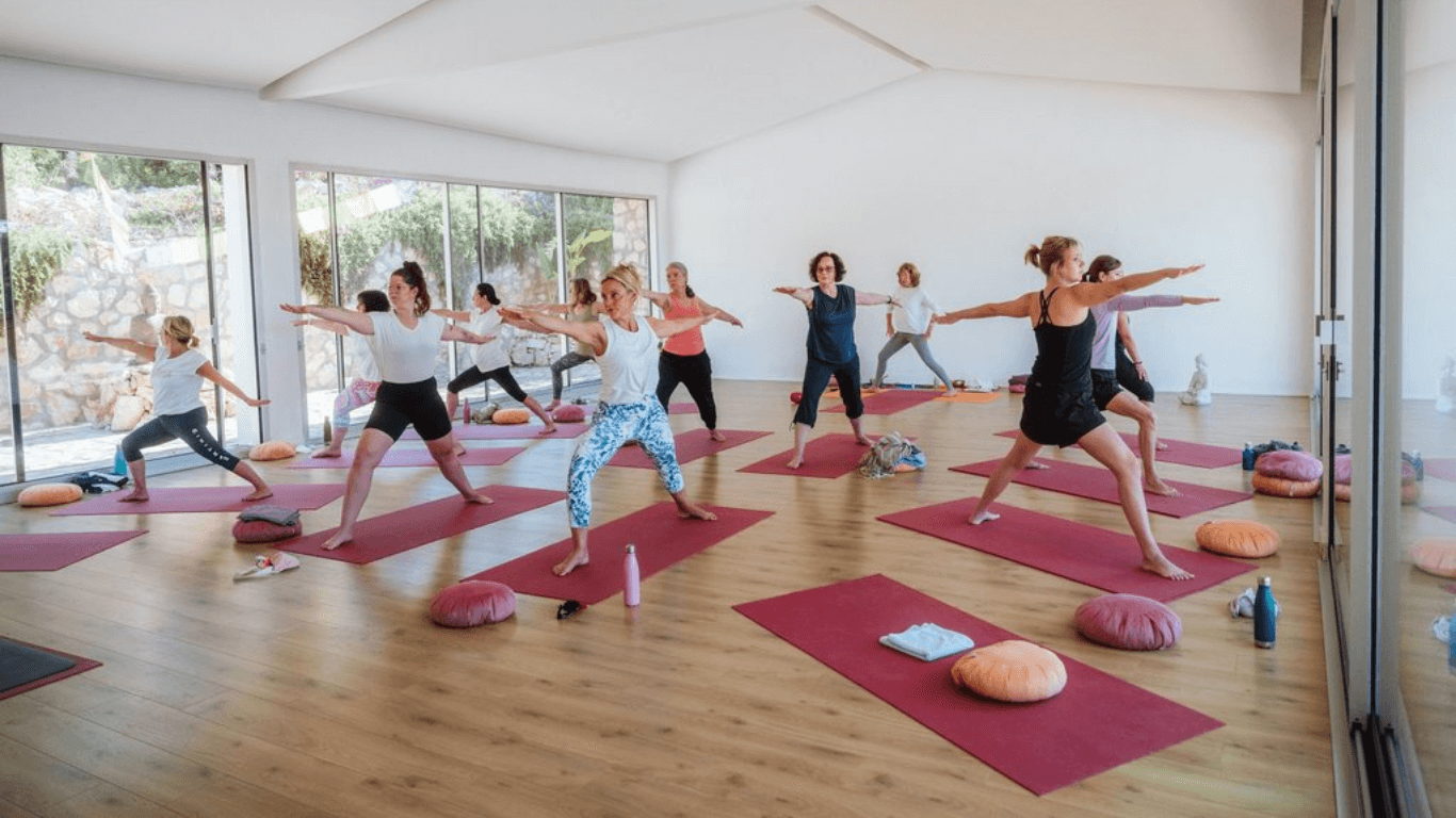 Eine Gruppe von Menschen macht Yoga in einem hellen, modernen Raum mit großen Fenstern. Sie stehen auf roten Yogamatten und lernen eine Yoga-Position