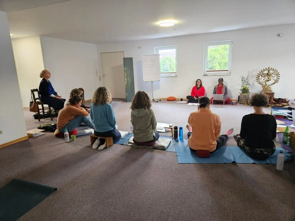 Gruppen von Menschen beim Yoga- oder Meditationskurs in einem hellen Raum, einige sitzen auf Matten im Schneidersitz, zwei Personen an einem Ende vor einem Whiteboard sind anscheinend die Lehrerinnen, es gibt eine Statue und eine Pflanze im Hintergrund.