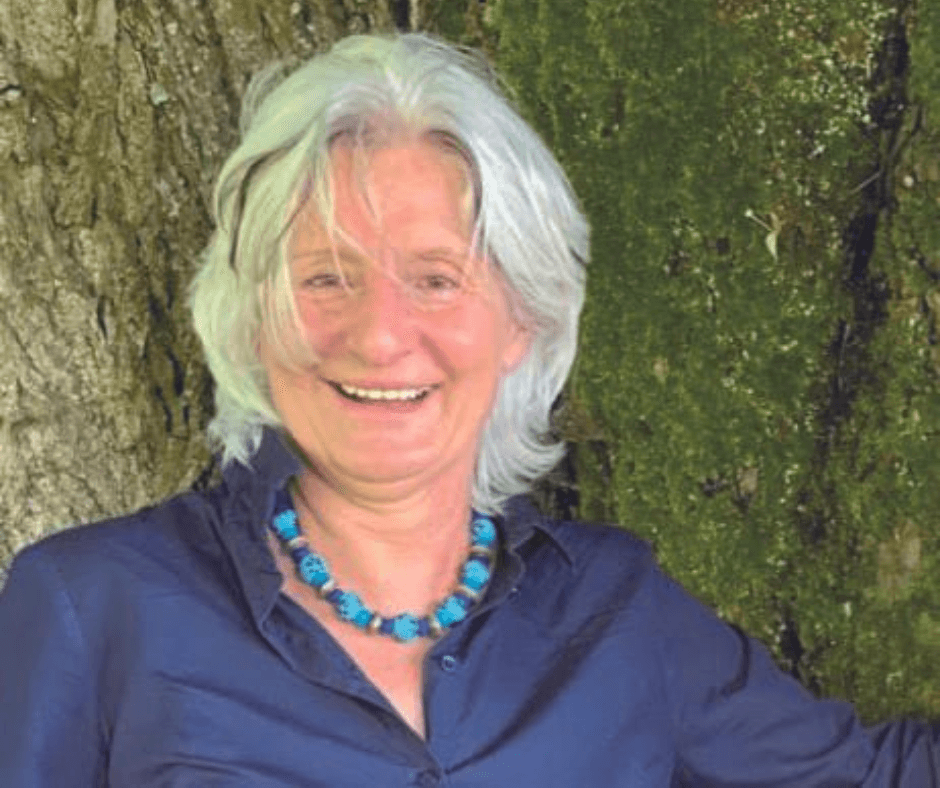 Anita Burkhart-Röser mit weißem Haar und blauem Kleid steht vor einem Baum im Freien und lächelt.