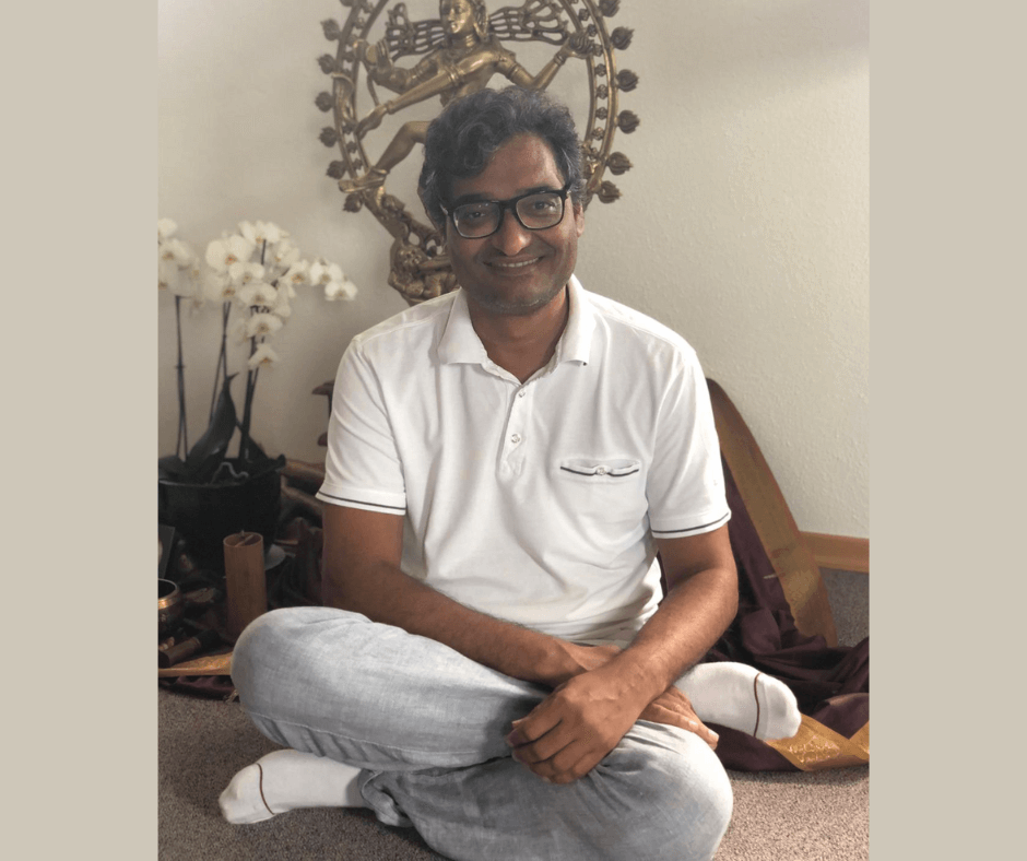 Anand Mishra in einer meditativen Sitzhaltung auf dem Boden, im Hintergrund eine Statue der hinduistischen Gottheit Shiva, umgeben von weißen Blumen. Er trägt ein weißes Polo-Shirt und graue Hosen.