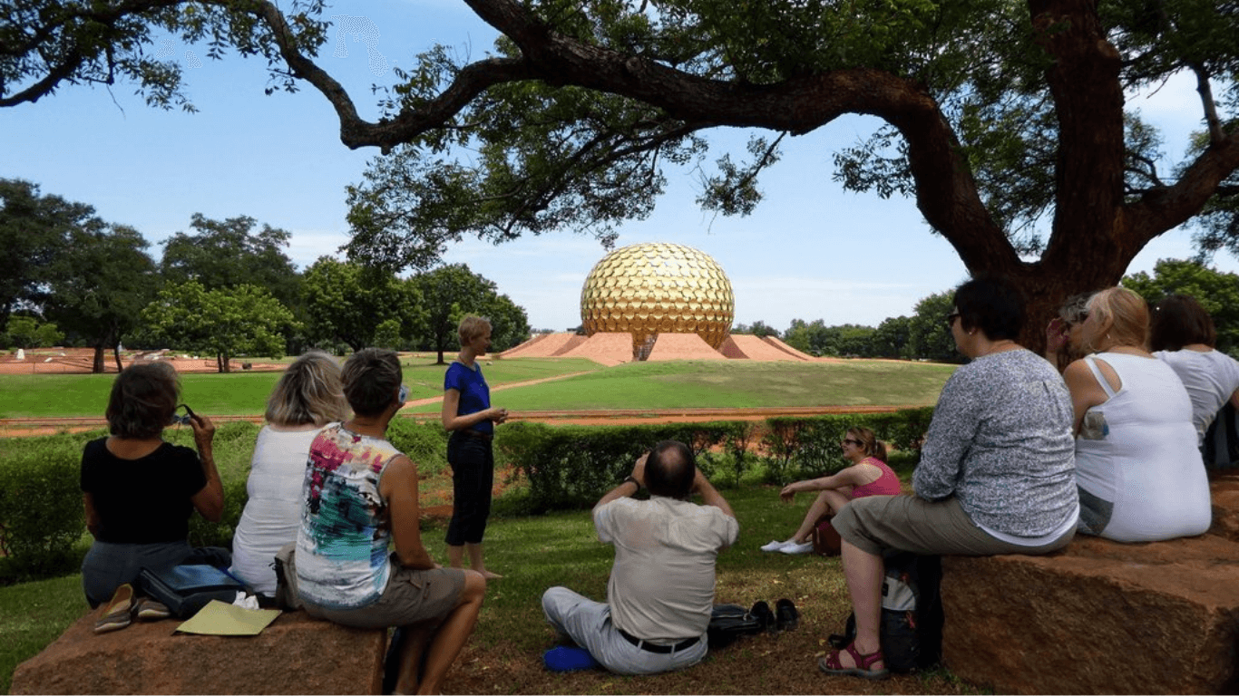Eine Gruppe von Menschen sitzt und steht im Park unter einem Baum und schaut auf ein großes, goldenes, kugelförmiges Bauwerk im Hintergrund.
