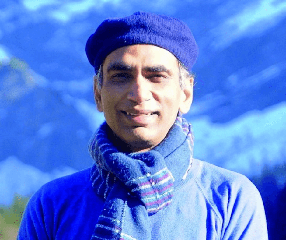 Mukesh Gupta trägt einen blauen Hut und Schal, im Hintergrund Berge und blauer Himmel.