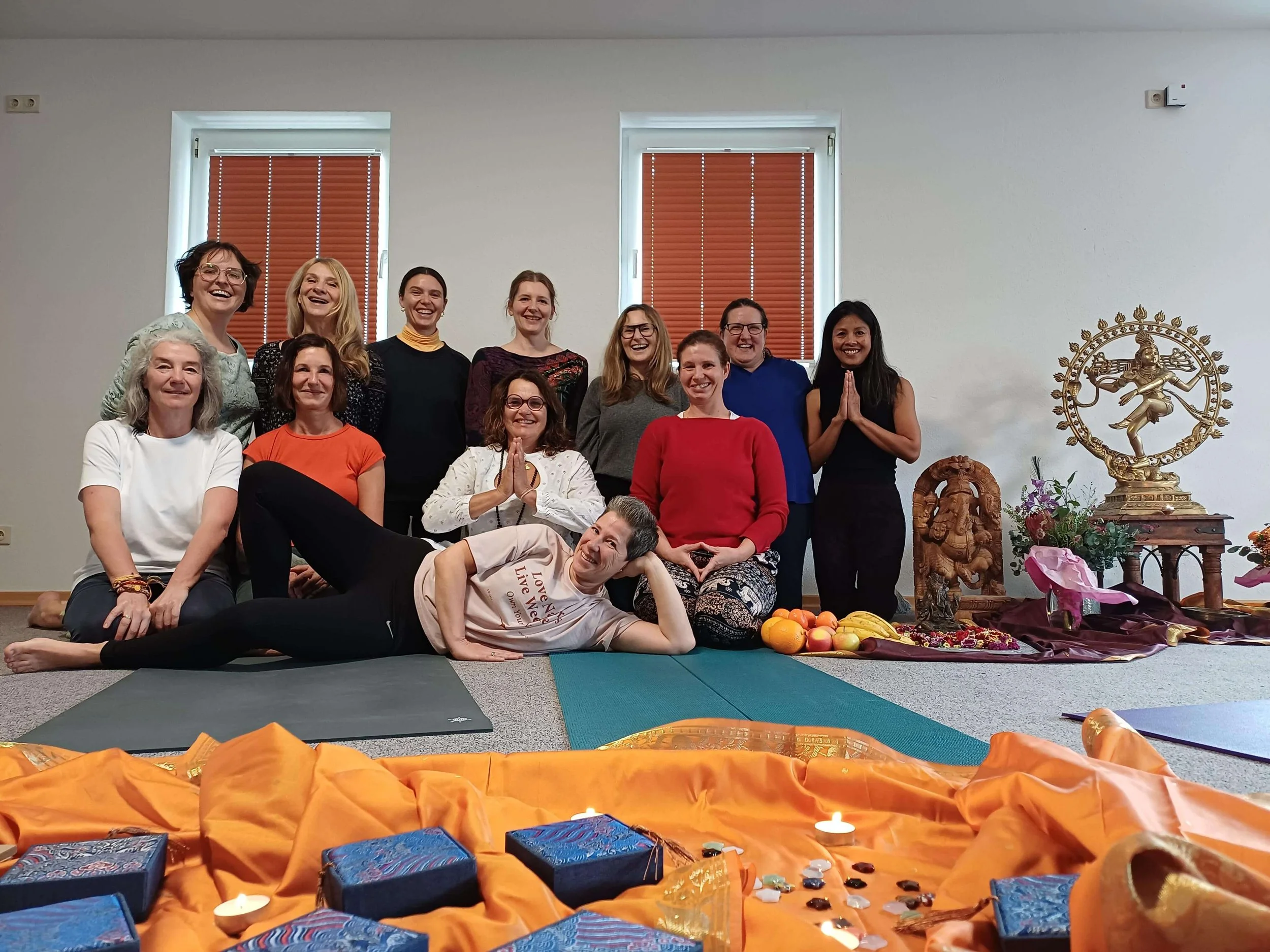 Abschlussgruppe Yogalehrausbildung BDY Okt.2025.  Sie lachen und posieren für die Kamera, einige haben die Hände im Gebet. Im Vordergrund sind Geschenke, Kerzen und Dekoration auf orangefarbenem Stoff sichtbar.