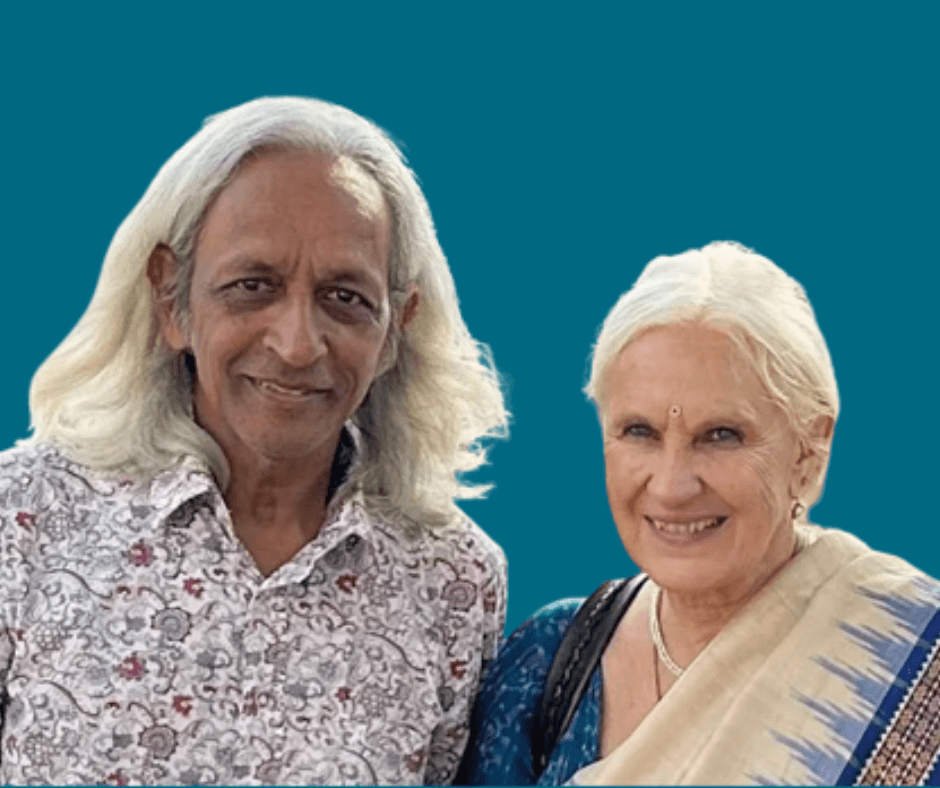R. Sriram und Anjali Sriram, ein Mann mit langen grauen Haaren und eine Frau mit kurzen weißen Haaren, stehen nebeneinander vor einem einfarbigen blauen Hintergrund. Sie lächeln freundlich.