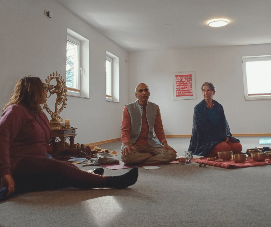 Drei Menschen sitzen auf dem Boden in einem hellen Raum, vermutlich bei einer Meditations- oder spirituellen Zeremonie, umgeben von Ritualgegenständen und einer goldenen Statue.
