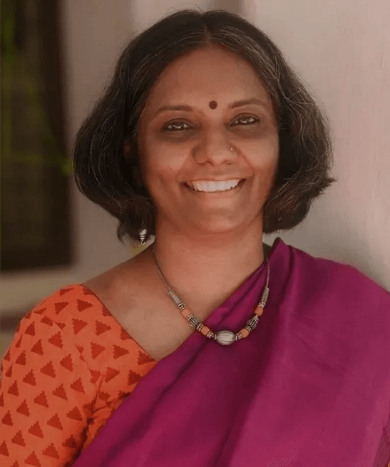 Sheela Shankar mit kurzen schwarzen Haaren, bindi auf der Stirn, trägt eine orange Kleidung mit Mustern und eine pinkfarbene Saree, sowie eine Halskette.