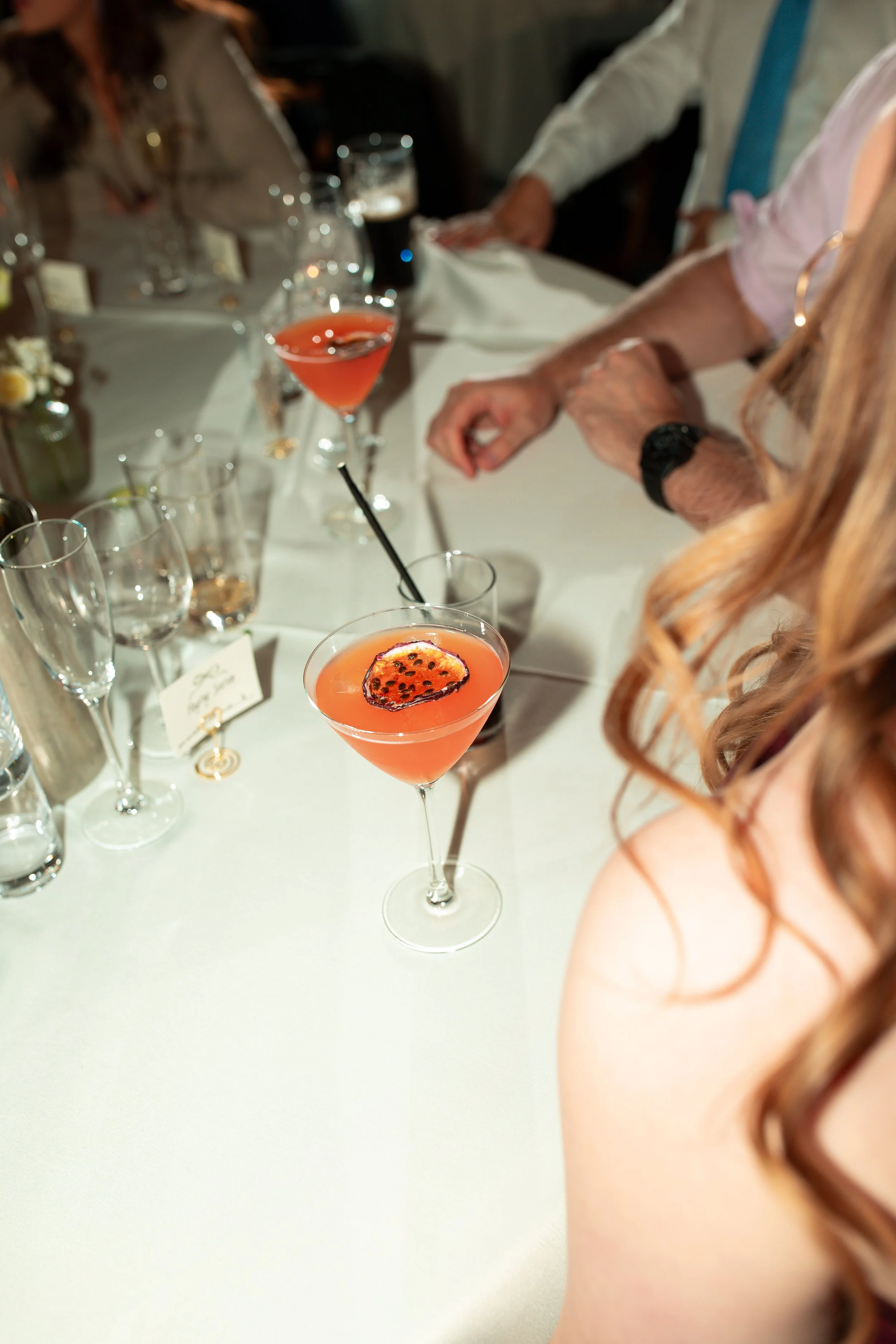At a formal dinner table, two pink cocktails garnished with passion fruit slices are in focus, with other empty glasses and a small flower arrangement in the background.