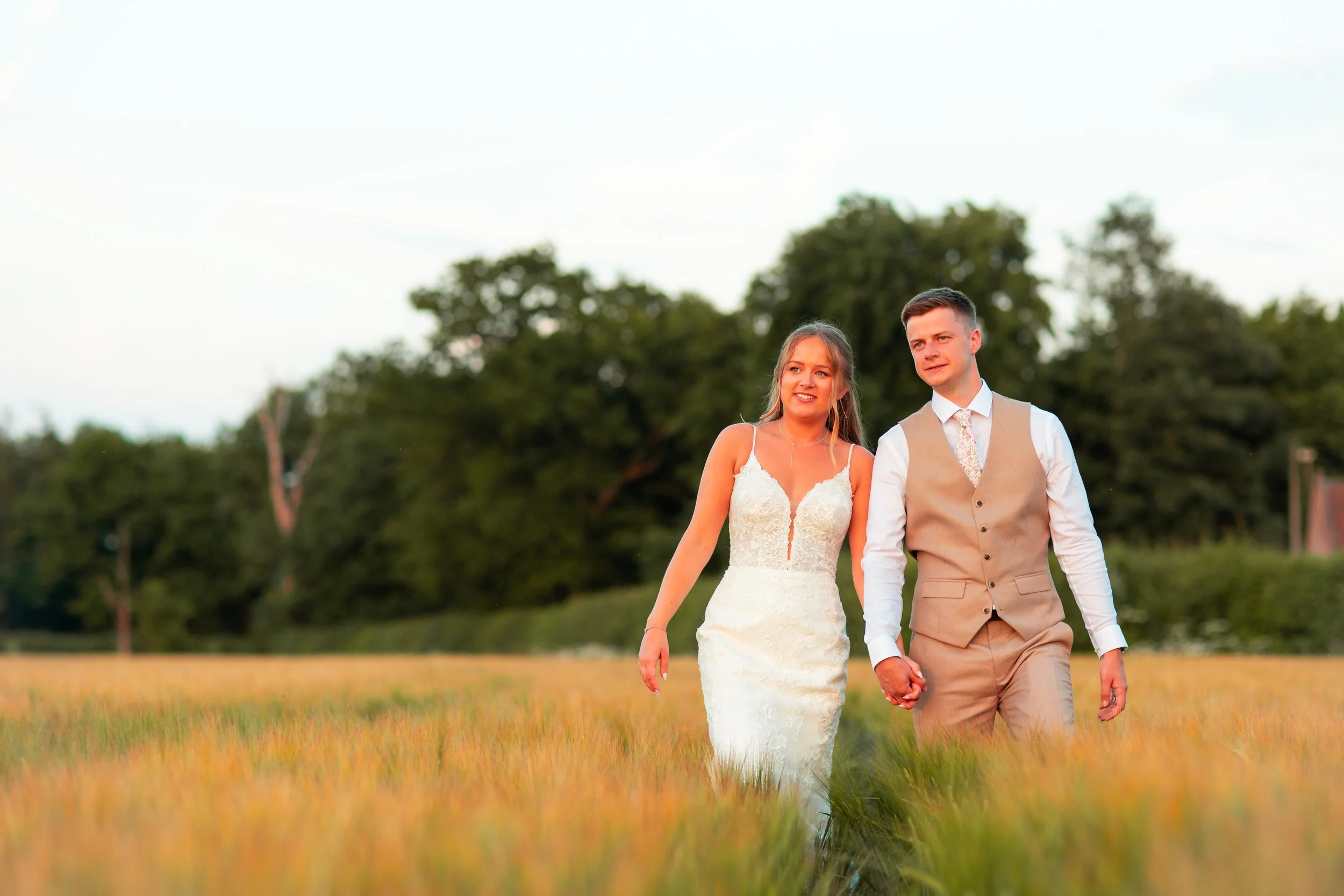 A bride and groom walking hand in hand through a field at sunset, dressed in wedding attire, with trees in the background.