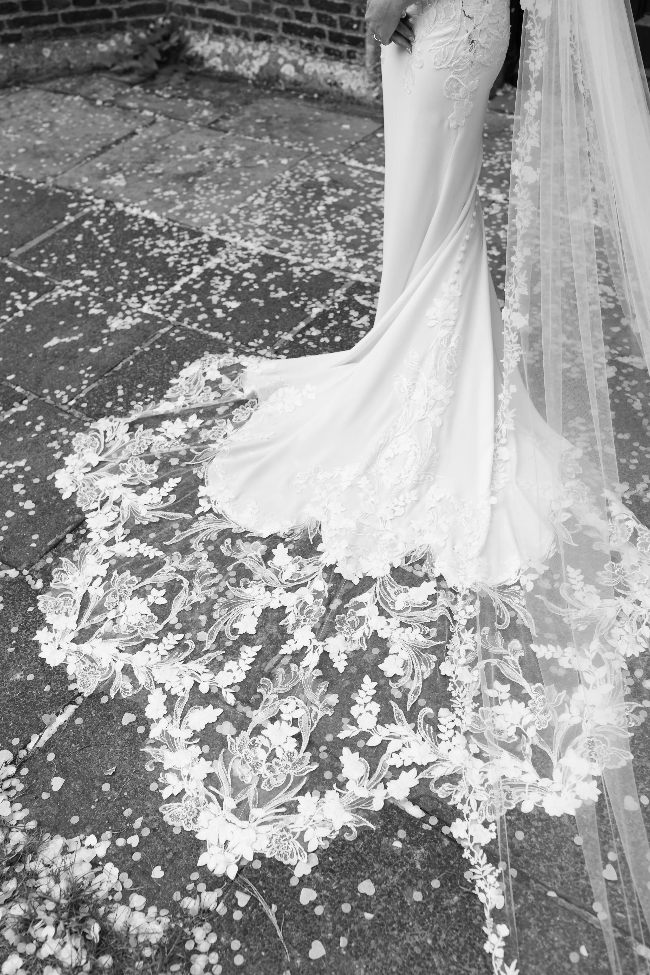 A bride in a white wedding gown with floral lace details stands on a stone pavement covered with scattered flower petals and a long lace veil trailing behind. The photo is in black and white.