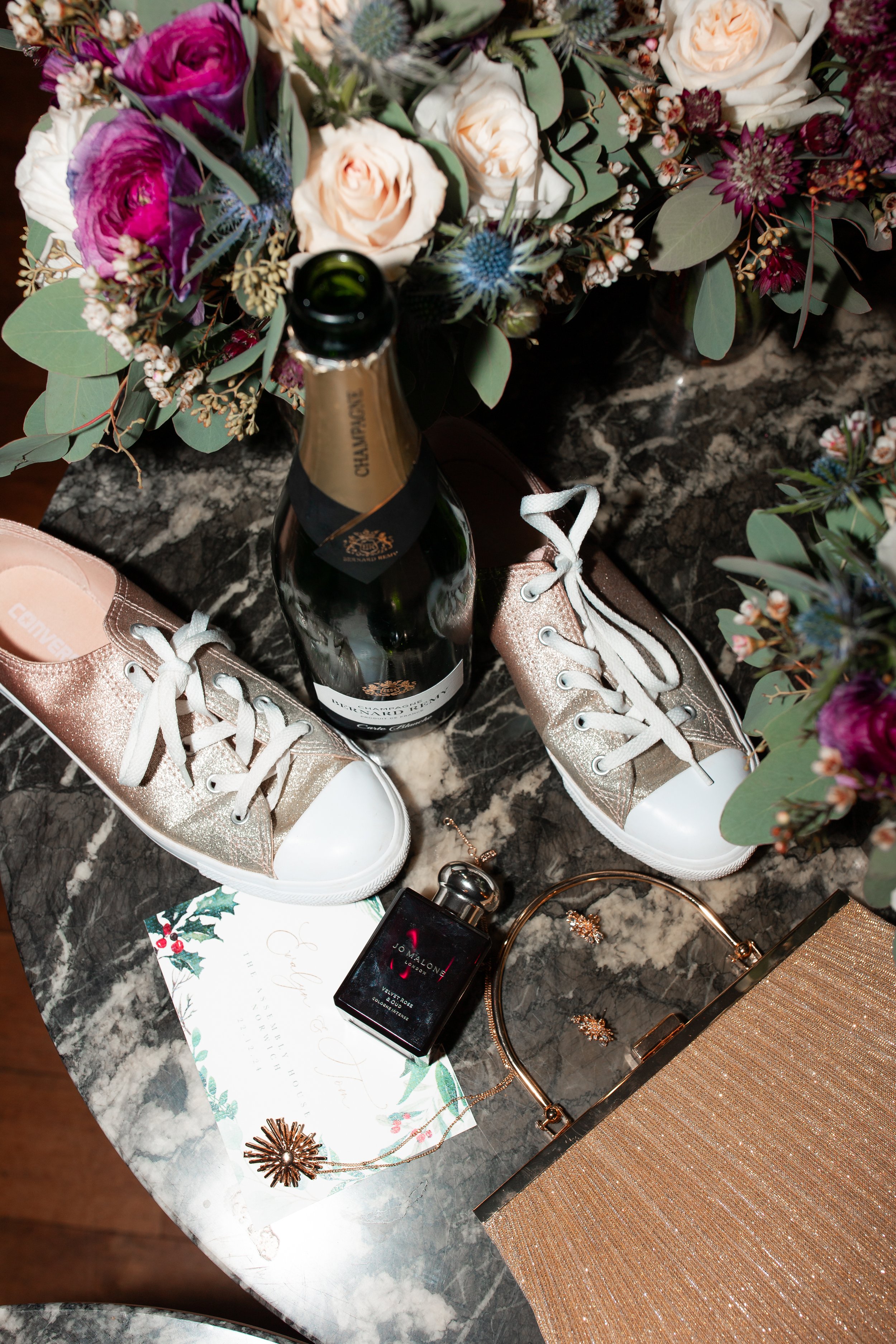 A marble tabletop with a large floral arrangement, a bottle of champagne, a pair of glittery gold sneakers, a bottle of perfume, a necklace, earrings, a clutch purse, and a holiday greeting card.