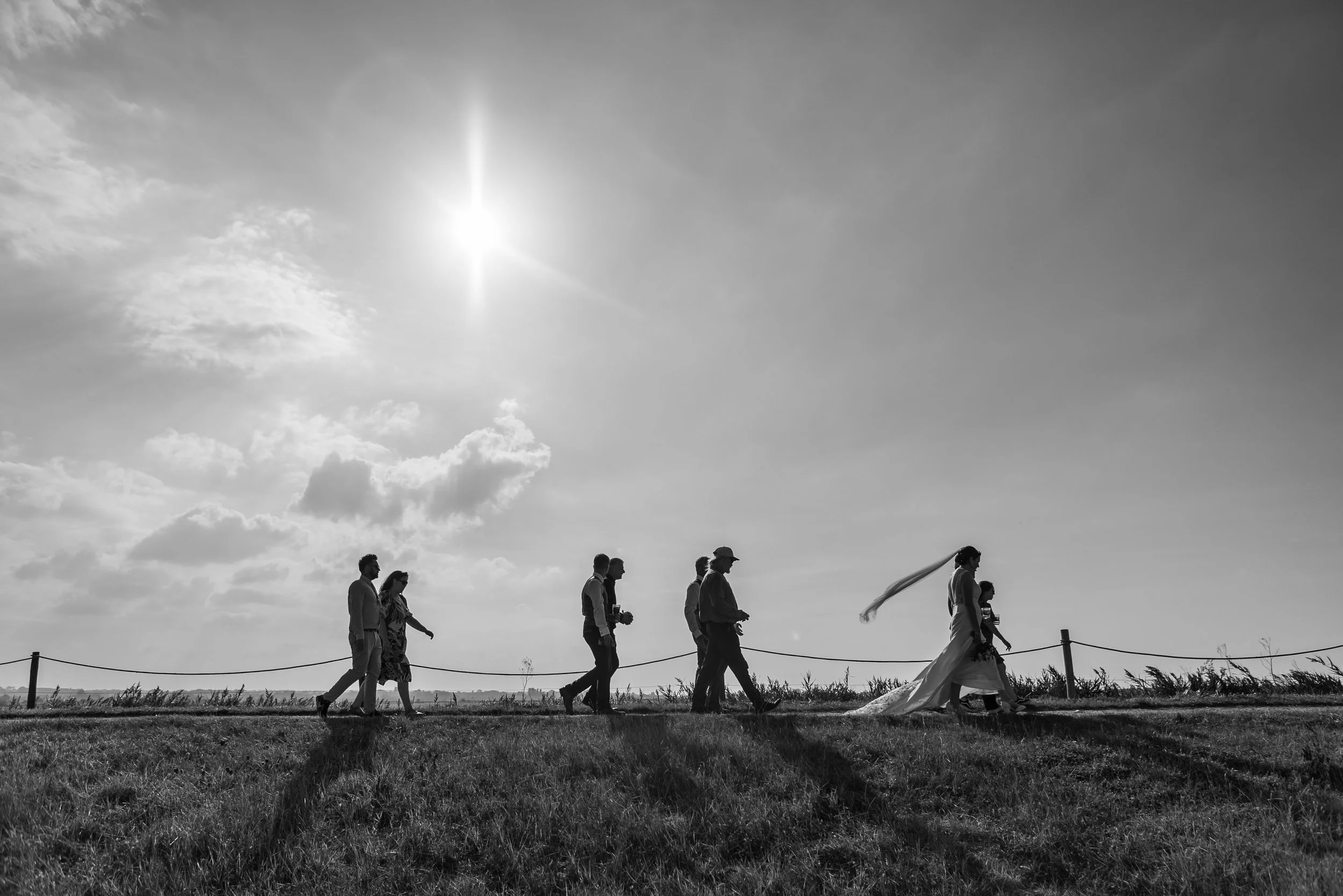 Black and white photo of a wedding procession walking outdoors on a grassy field under the sun.