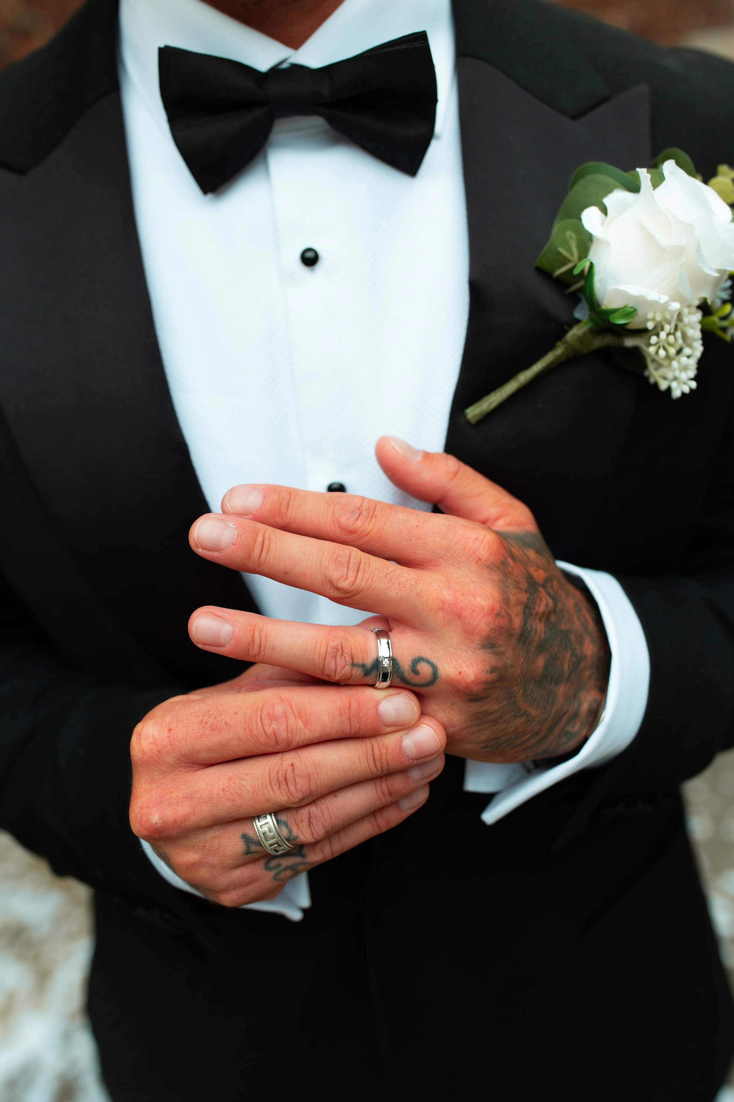Close-up of a man in a black tuxedo with a white shirt and black bow tie, showing his tattooed hands with wedding rings, and a white boutonniere on the lapel.