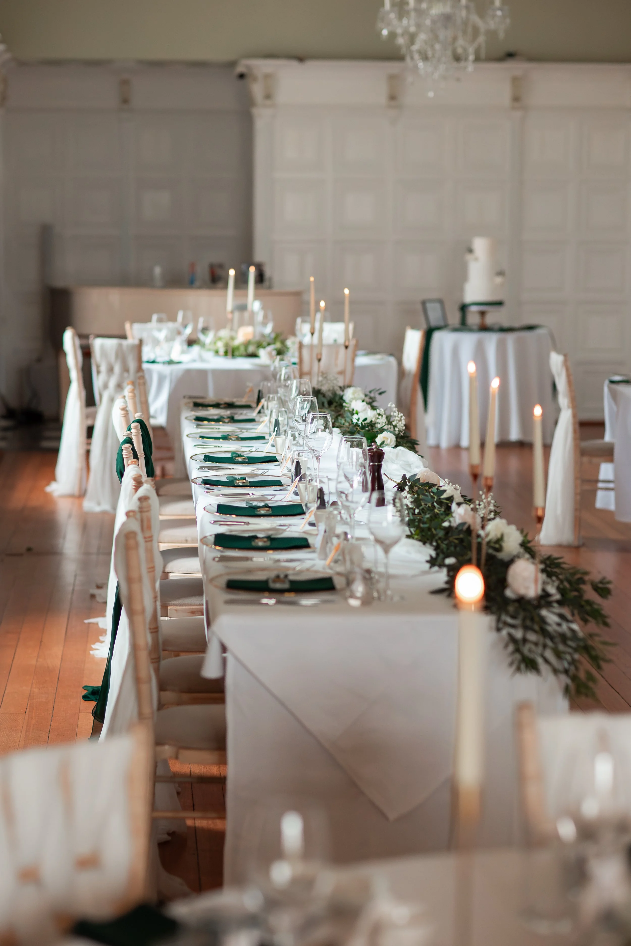 Elegant wedding reception table decorated with white tablecloths, green napkins, glassware, candles, and floral arrangements with white flowers and greenery, set in a bright, classic room with a chandelier.