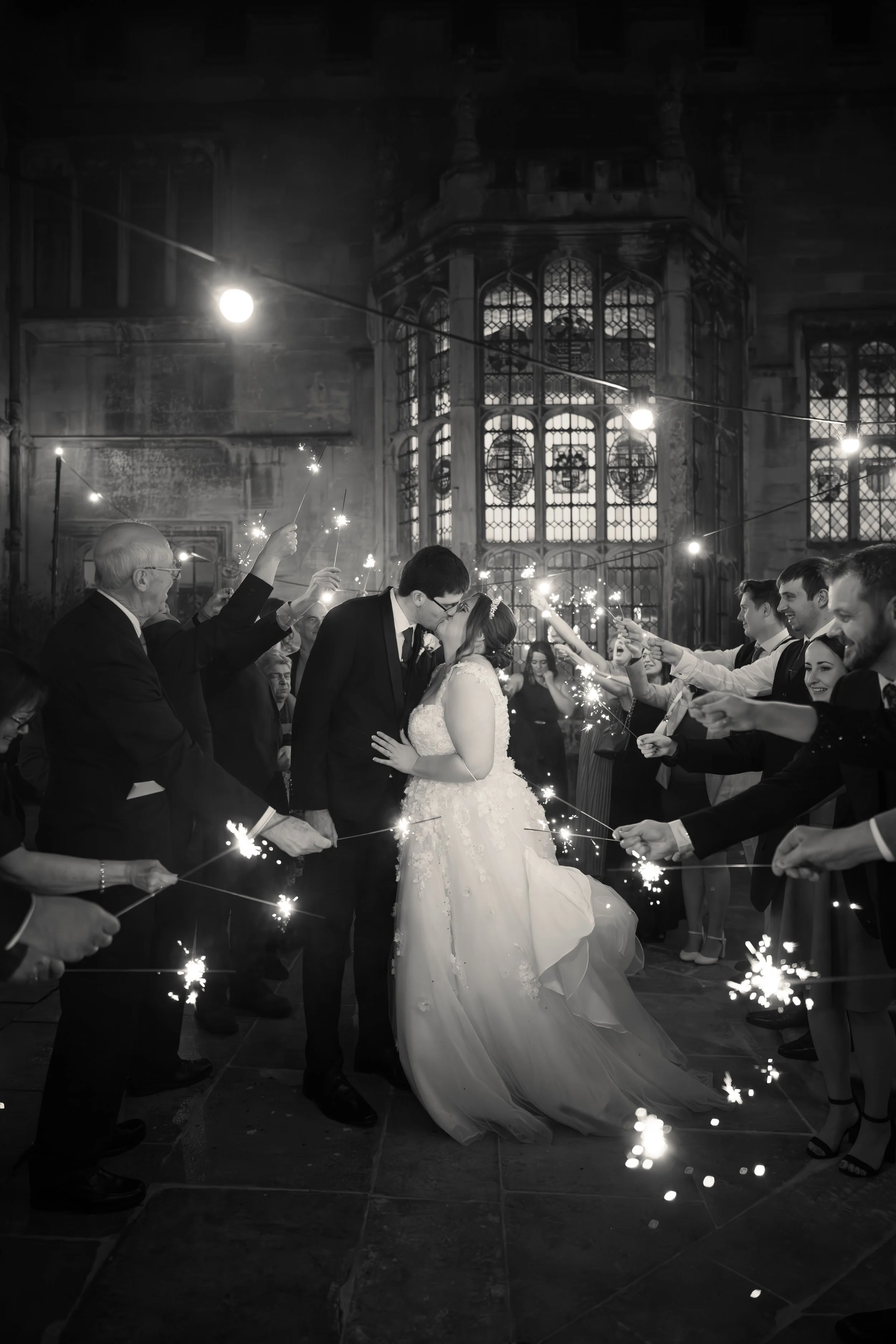 A black and white photo of a wedding celebration showing a bride and groom kissing in the center, surrounded by people holding sparklers inside a grand church with large stained glass windows.