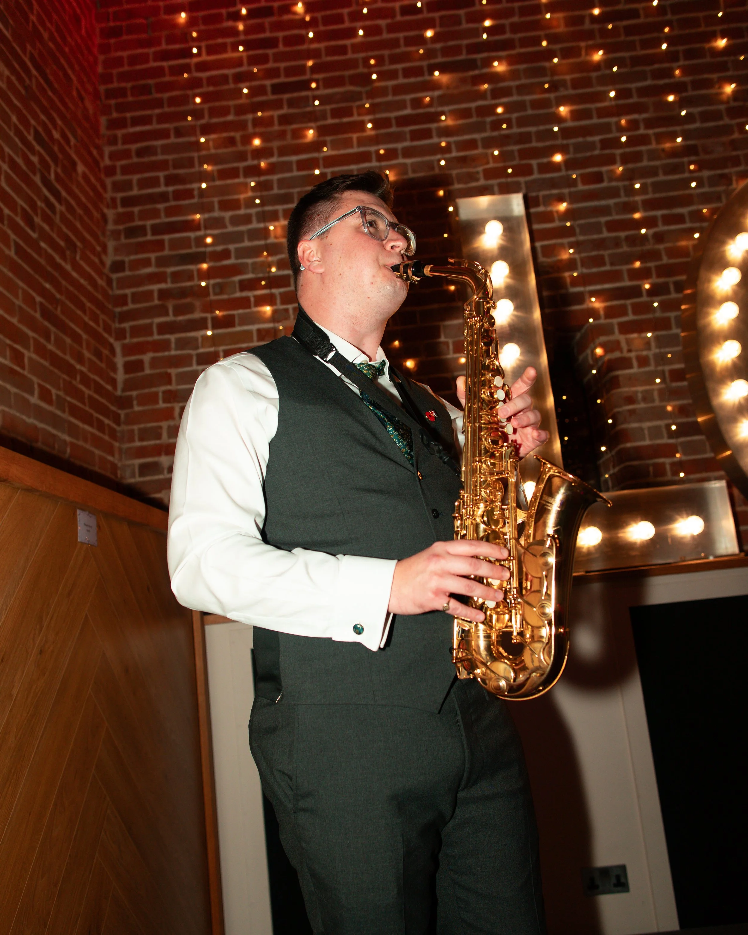 A man in business attire playing a saxophone at an indoor event, brick wall decorated with string lights in the background.