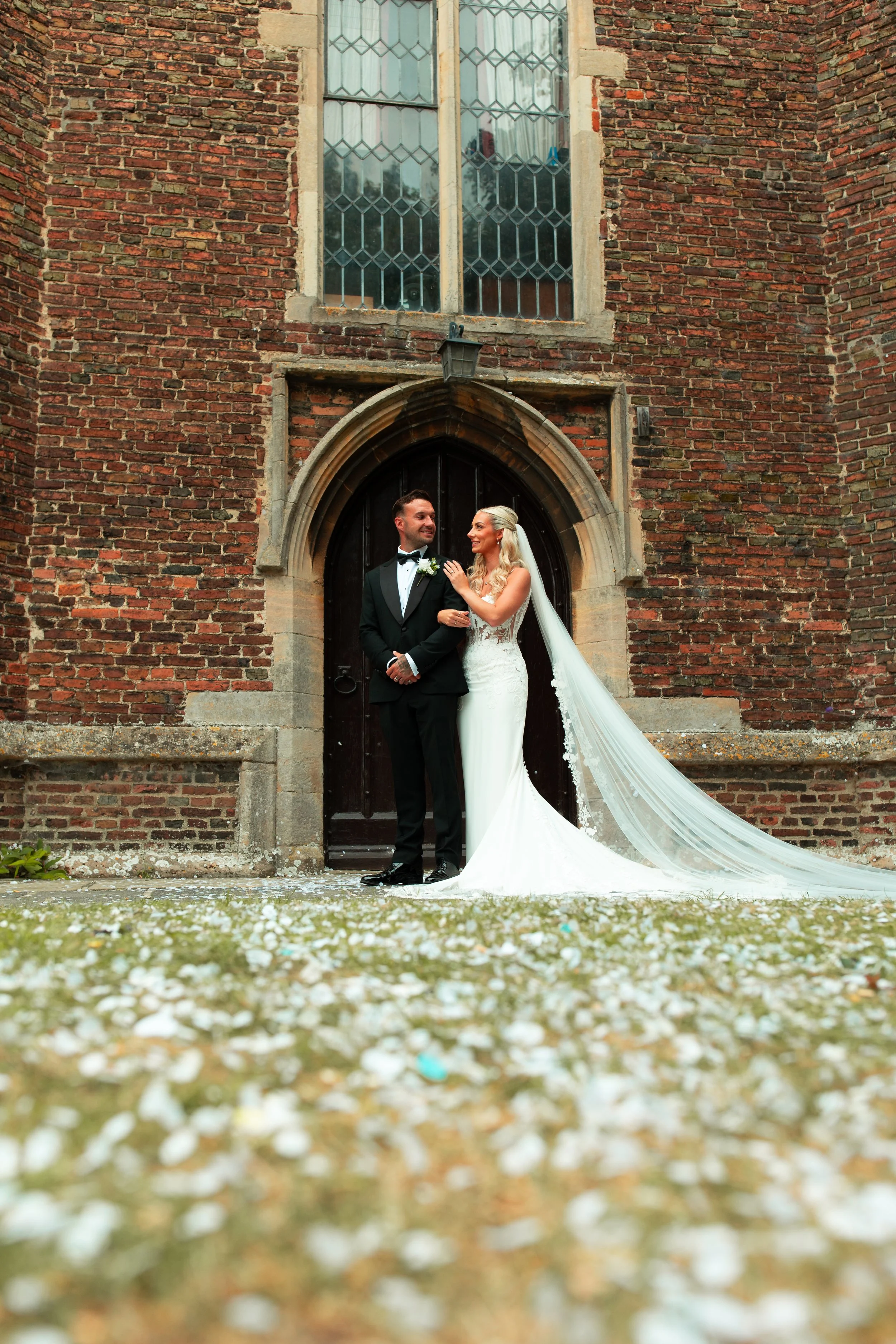 Bride and groom smiling and looking at each other in front of a brick church wall with large window, wedding flowers on ground.