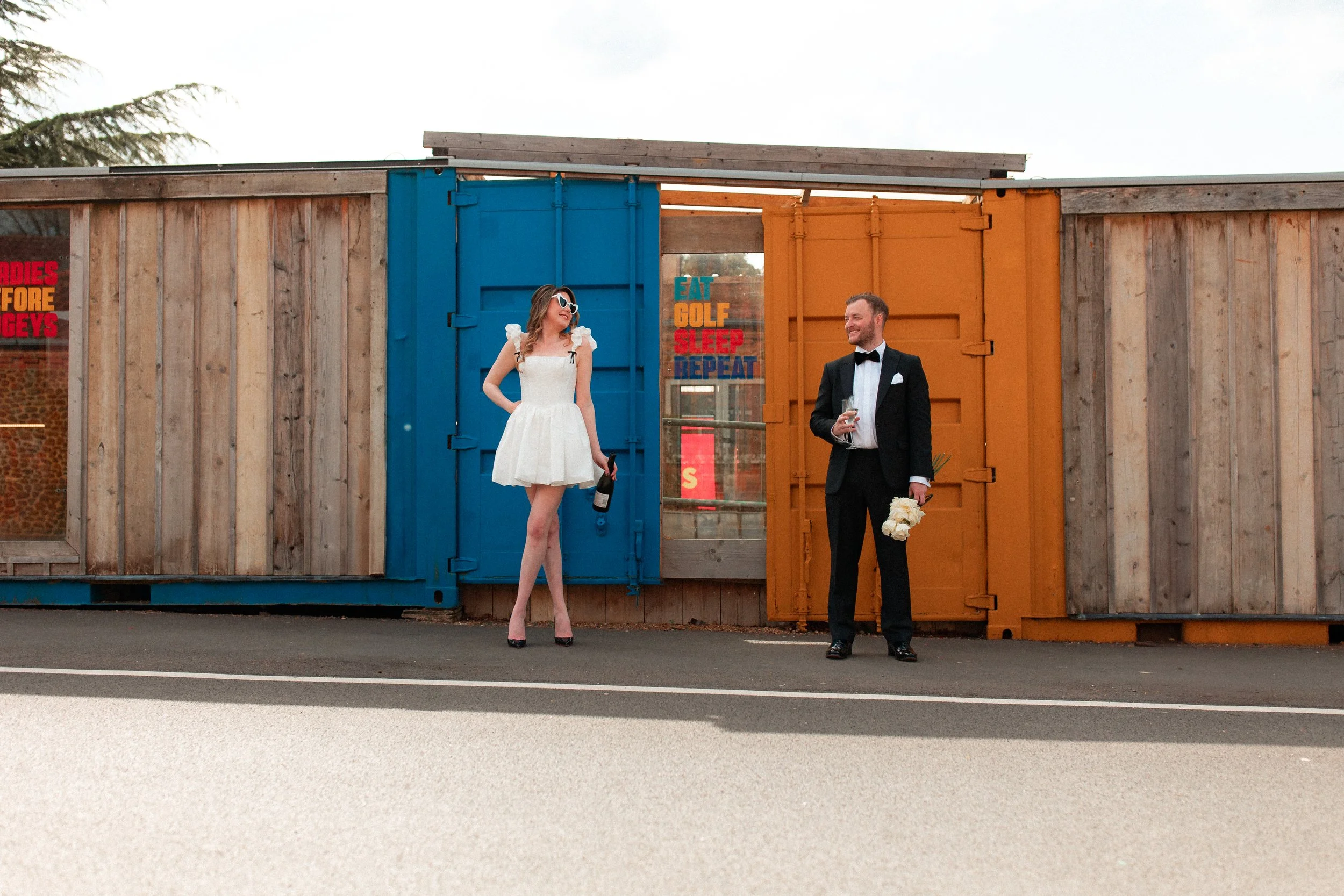 A woman in a white dress and sunglasses standing next to a man in a tuxedo holding a bouquet and a glass of wine in front of colorful shipping containers.