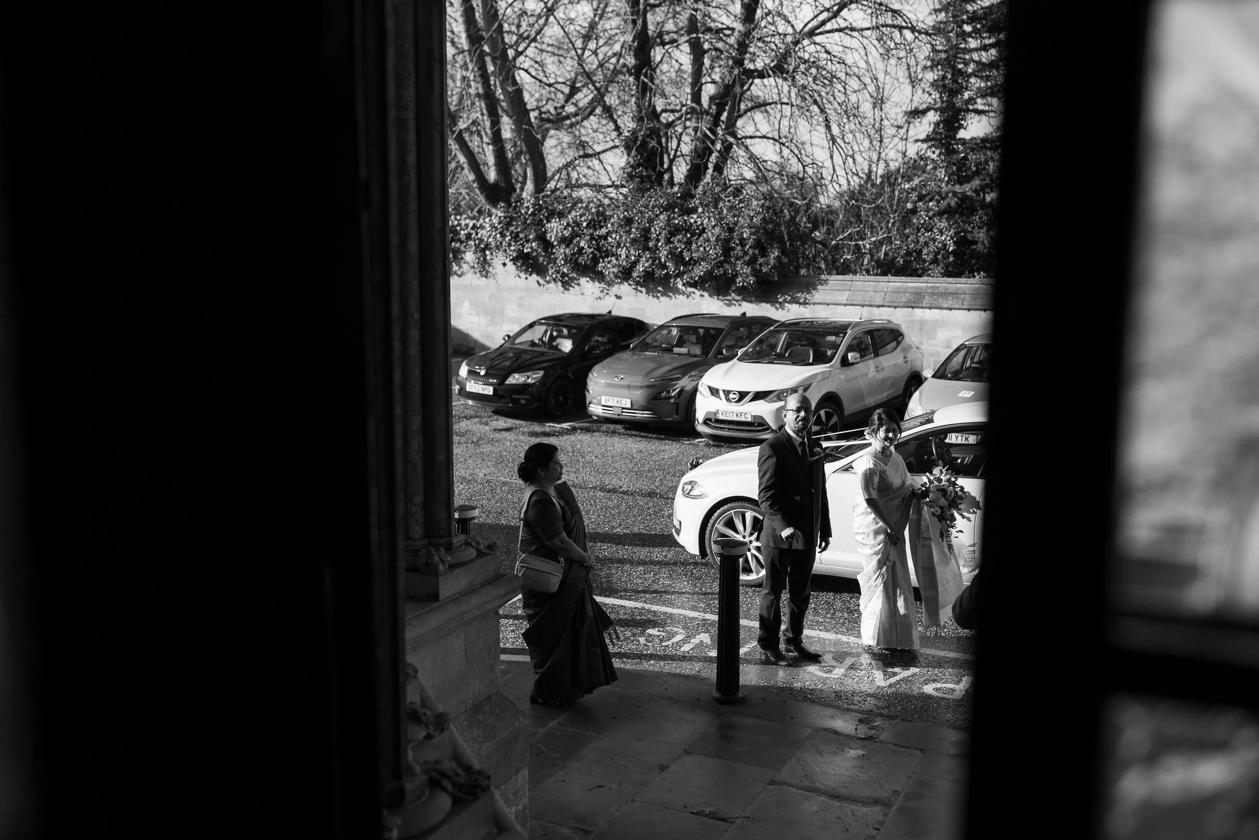 Black-and-white photo of a woman in a sari holding a bouquet, a man in a suit, and another woman walking on a street outside a building with parked cars in the background.