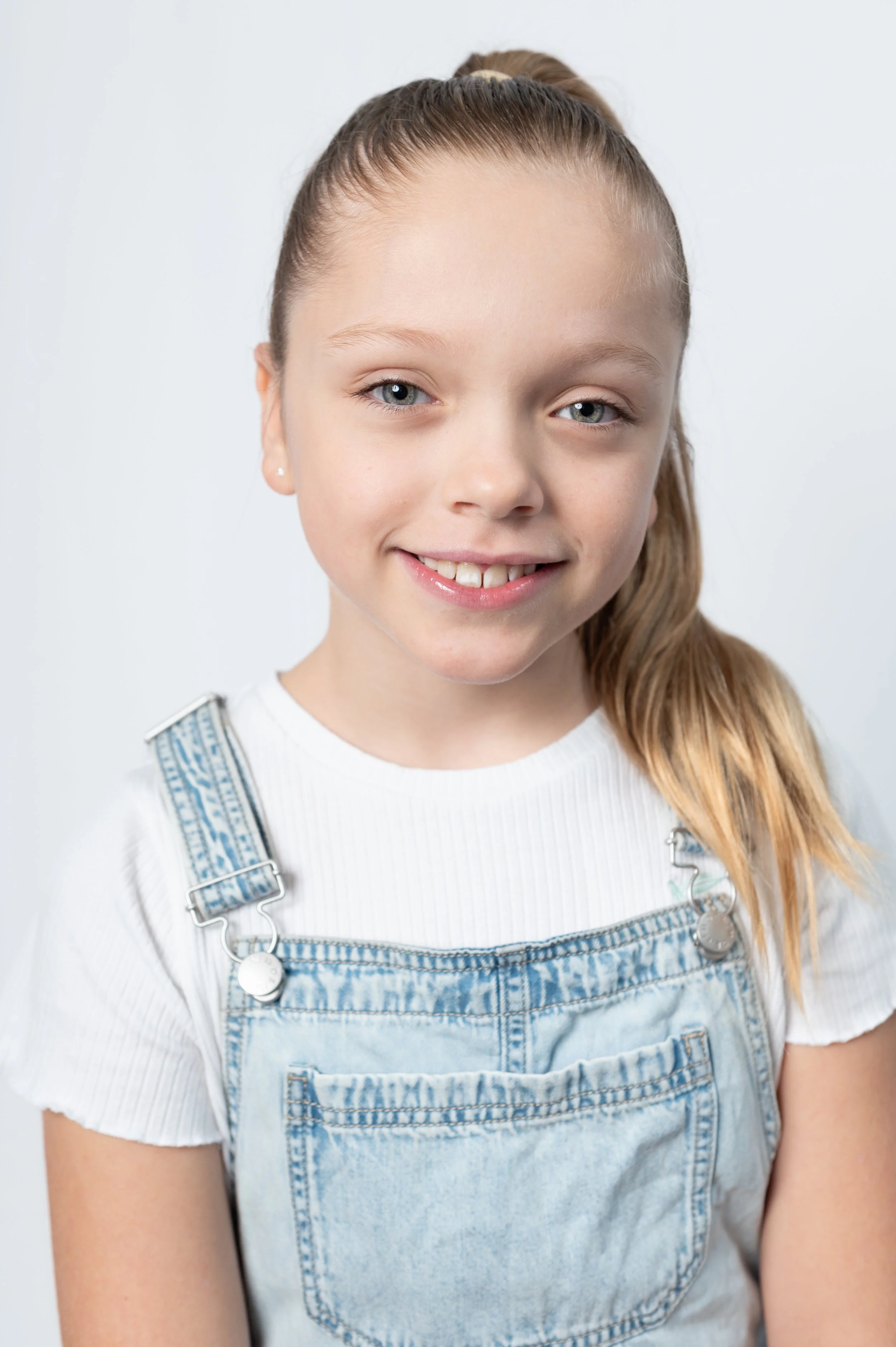 A young girl with blonde hair tied back in a ponytail, smiling, wearing a white t-shirt and light blue denim overalls, standing against a plain light background.
