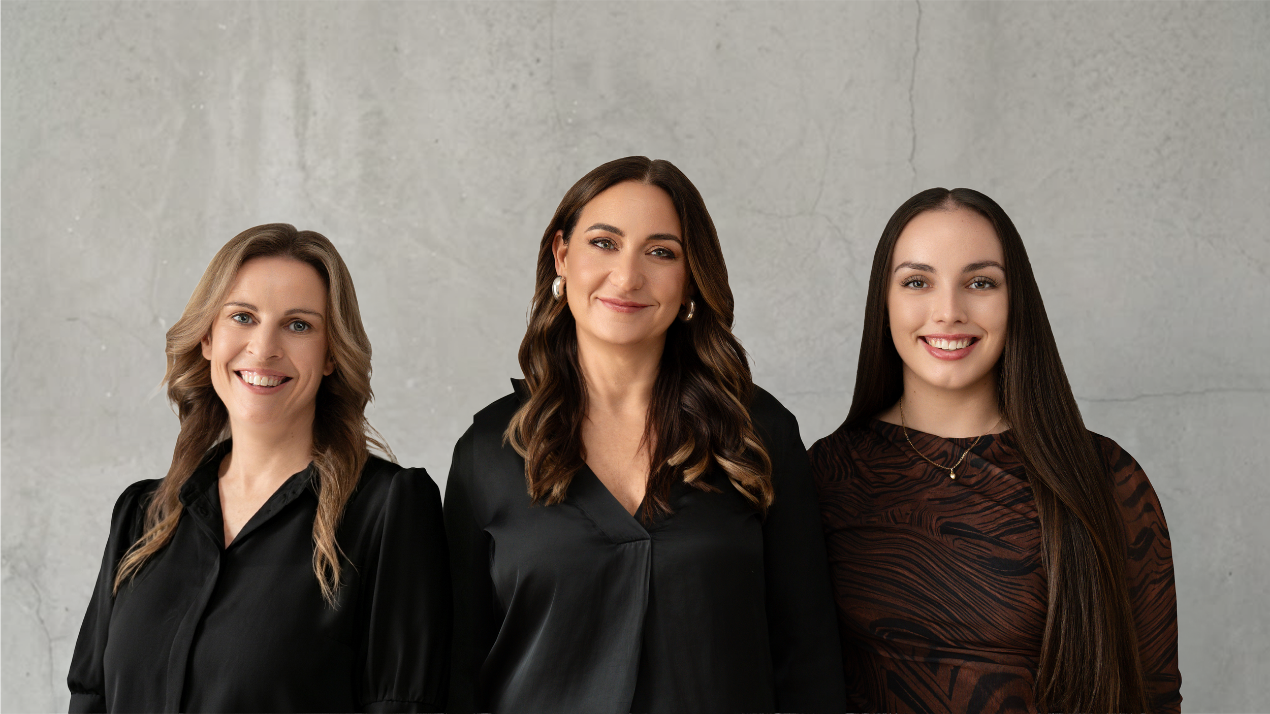 Three women standing side by side posing for a photo against a light gray wall, smiling, dressed in dark clothing.