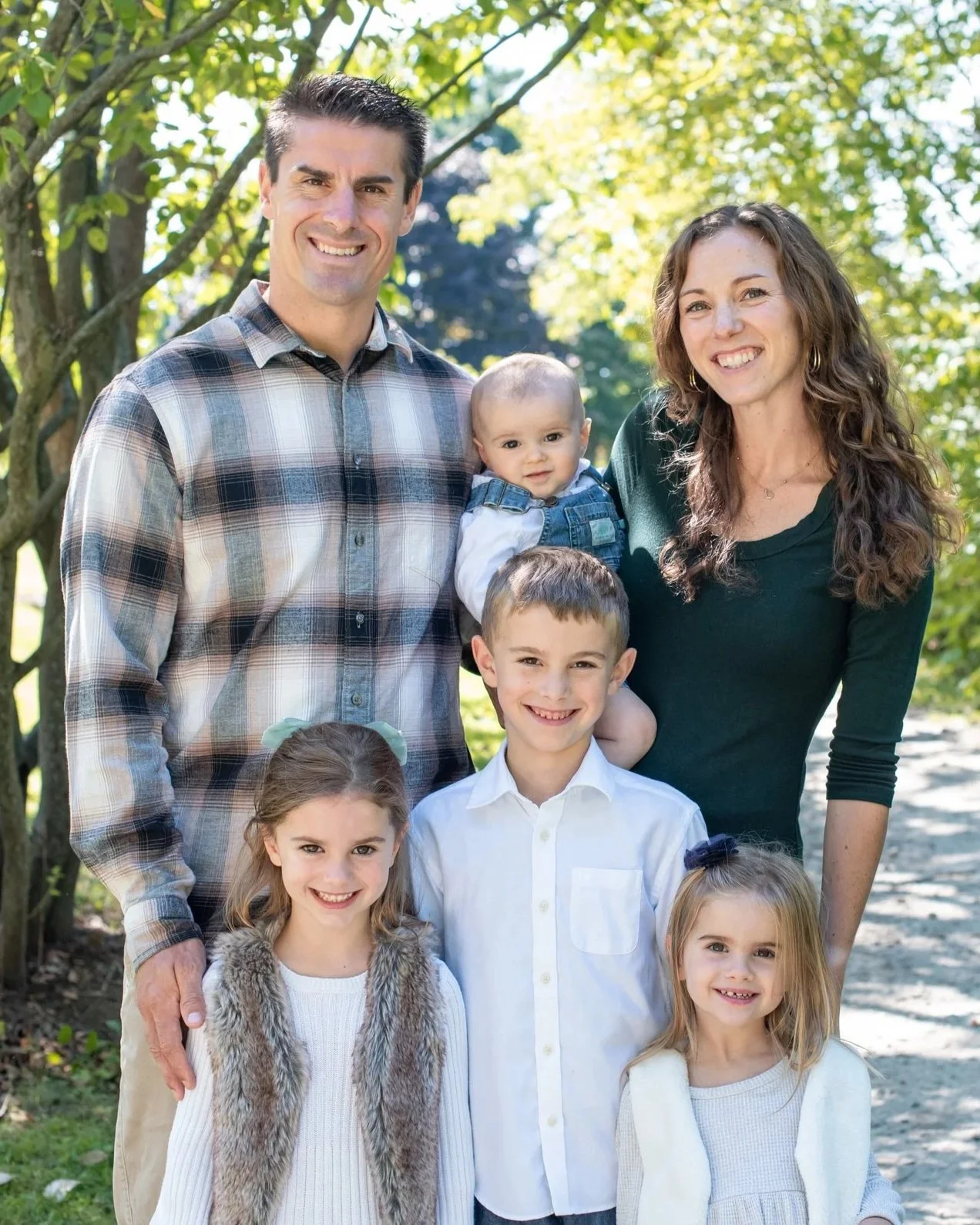 A family of seven posing outdoors on a sunny day, standing among trees with green leaves.