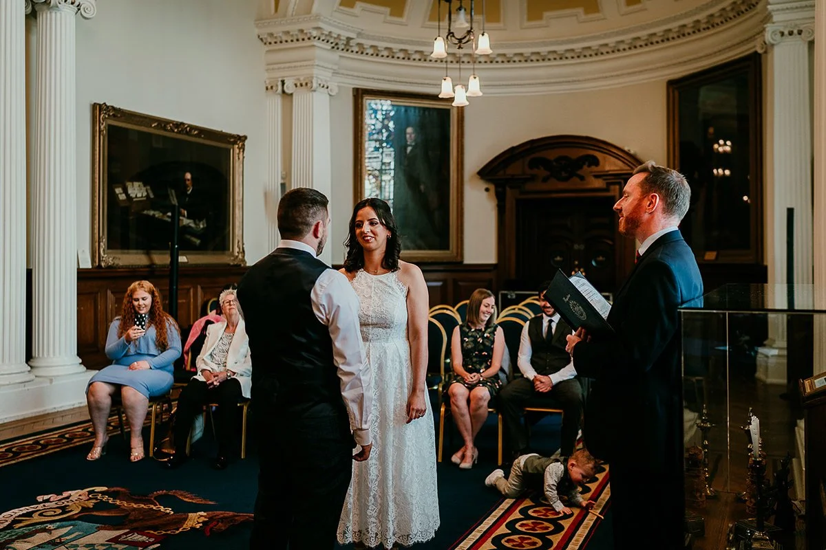 bride & groom exchanging wedding vows in Belfast City Hall. Bride facing groom & view of back of groom with guests visual in the background