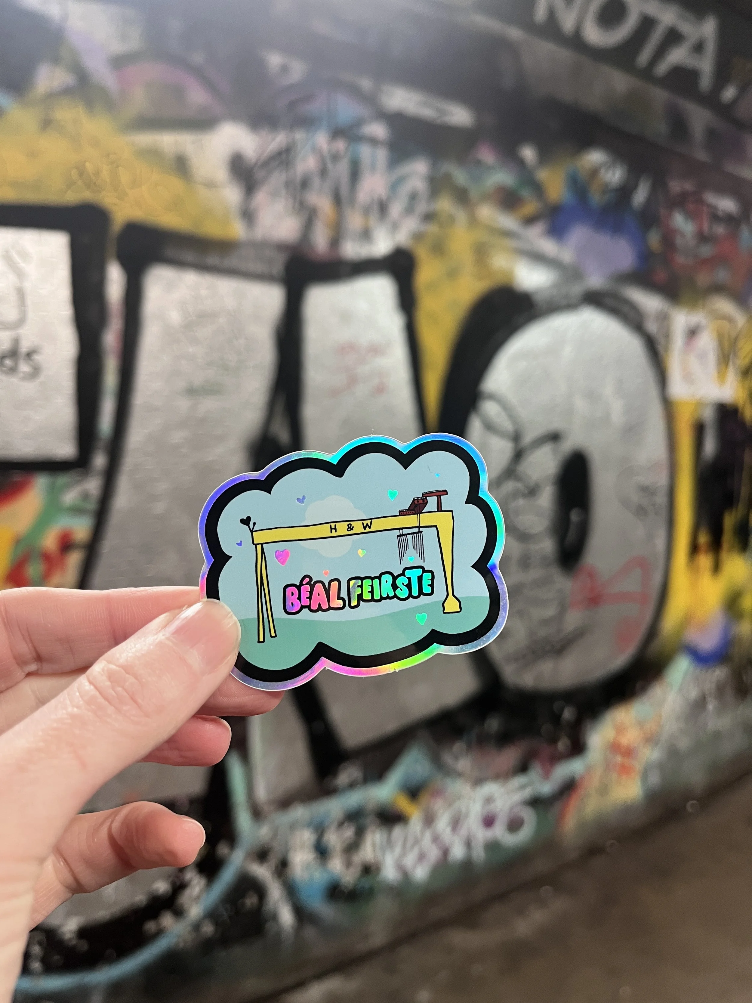 A hand holding a holographic sticker with colorful text and graphics in front of a graffiti-covered wall. The sticker features a crane and the words "Béal Feirste." Irish Language sticker in a Belfast underpass.