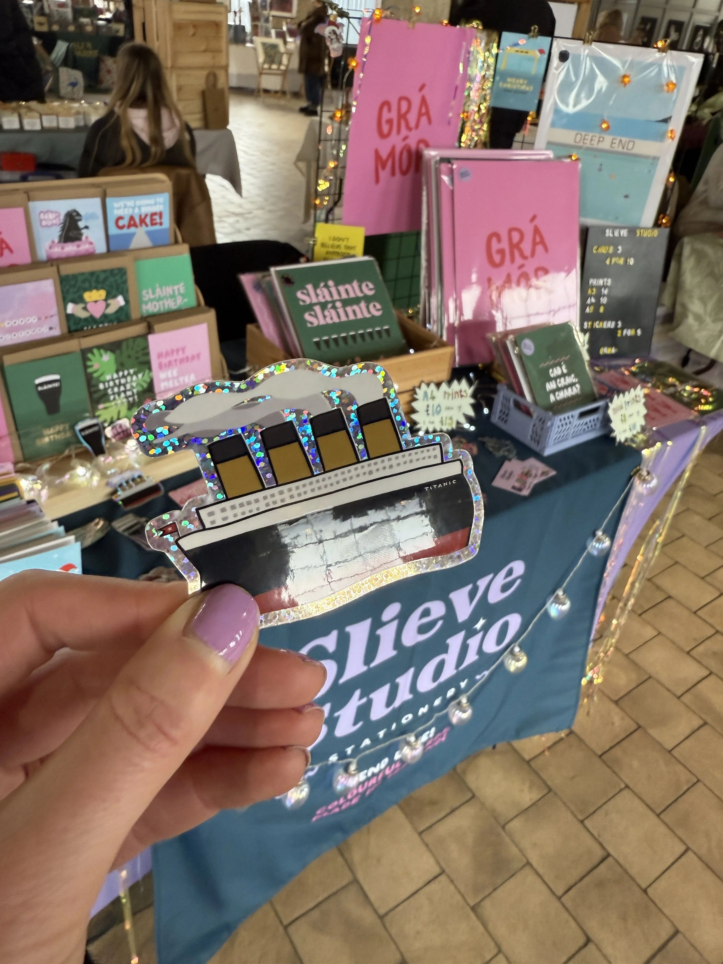 Photograph of a hand holding a shiny sticker of the Titanic ship with its smokestacks. In the background, a booth displays various colorful books and items for sale, with signs and fairy lights decorating the stall.