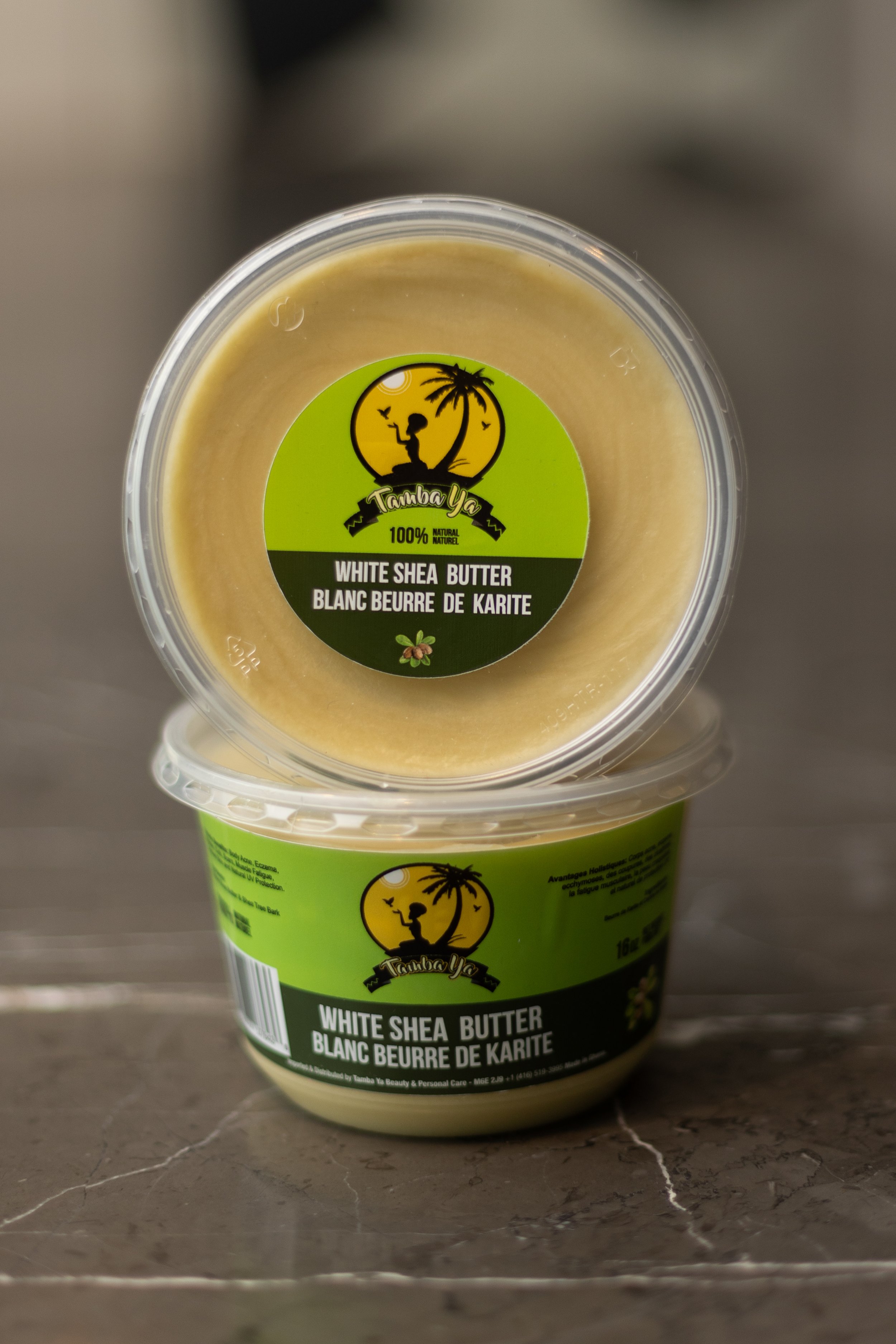 Two containers of Tamba Ya white shea butter stacked on a gray surface. The top container is viewed from above, showing the label with a logo of a person sitting under a palm tree with birds, and the text 'White Shea Butter' in English and French.