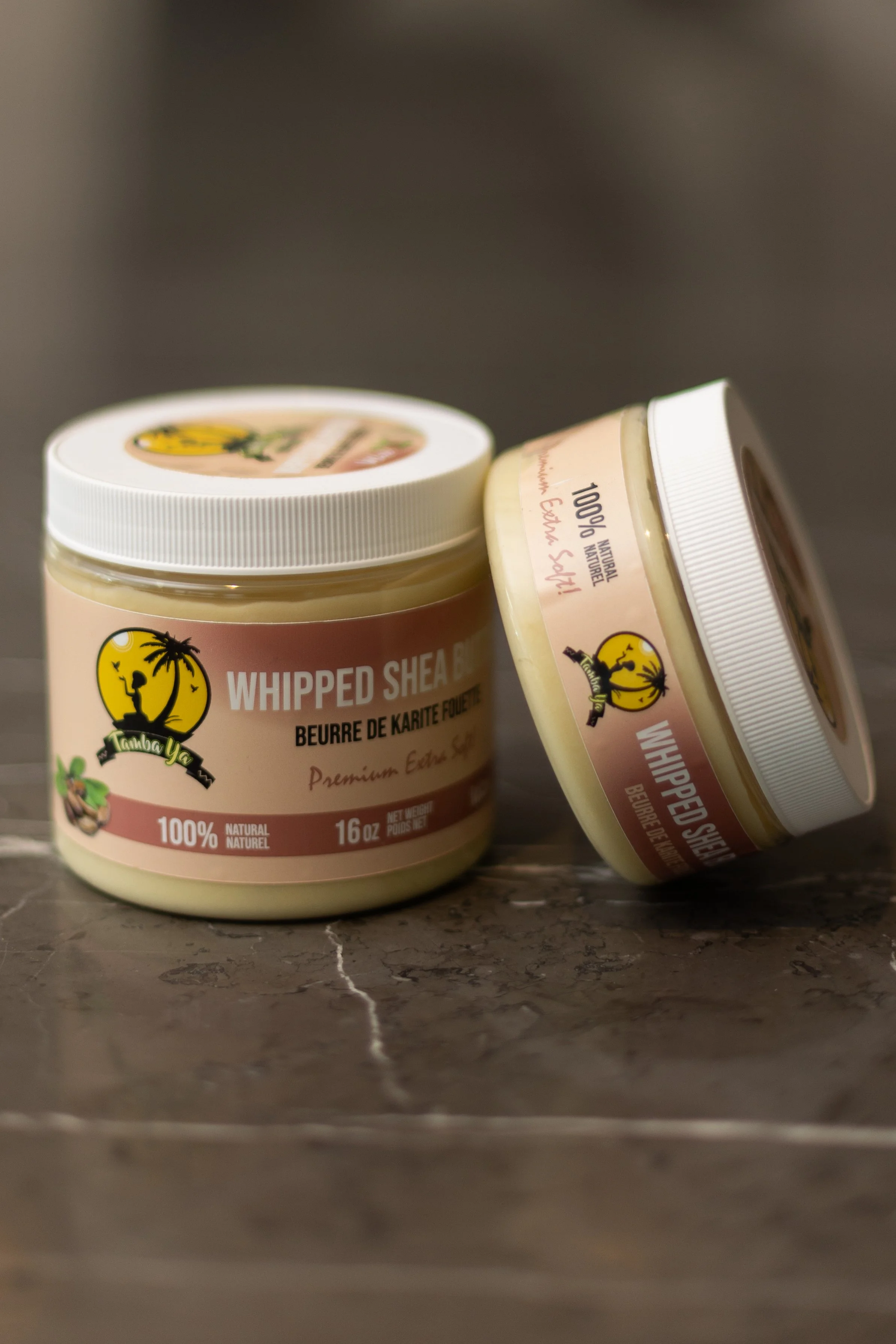 Two jars of whipped shea butter on a marble surface, with the label showing a yellow circle and a silhouette of a person under a palm tree.