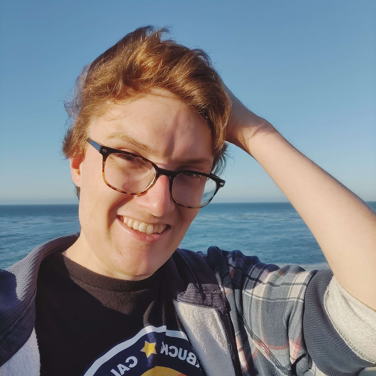 close up of a person with short red hair and glasses standing outside with the ocean behind them