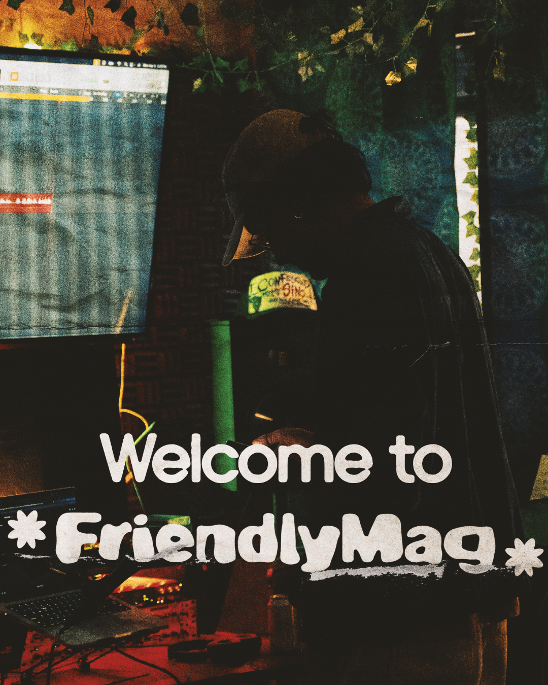 A person working on a laptop at a desk-themed event called Friendly Mag, with a sign saying 'Welcome to Friendly Mag' and a map of the world on the wall. The person is wearing a cap and dark clothing, and there are computer equipment and decorations around.