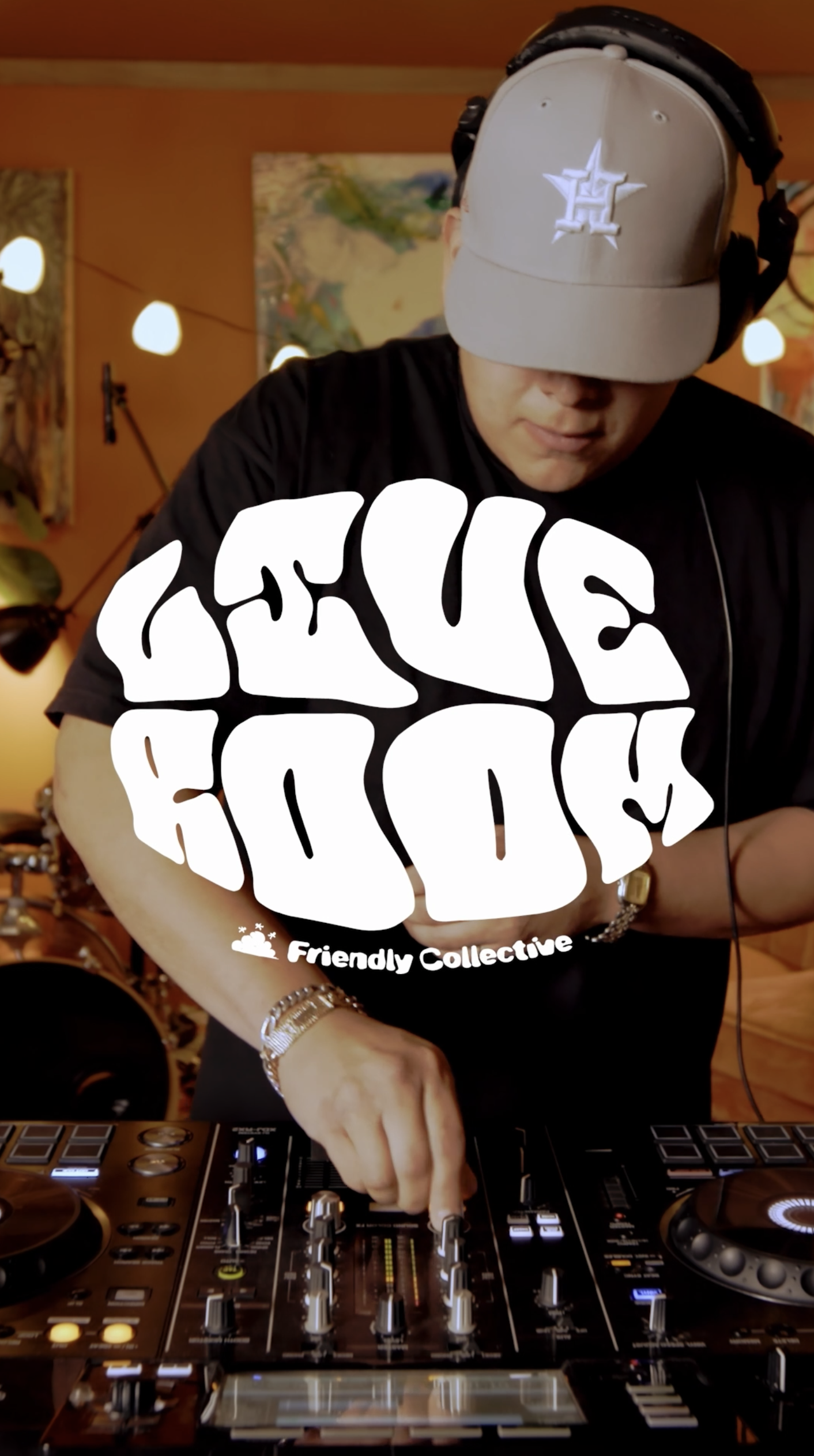 A DJ wearing a baseball cap and headphones, working with a DJ mixer in a warmly lit room decorated with art and string lights.