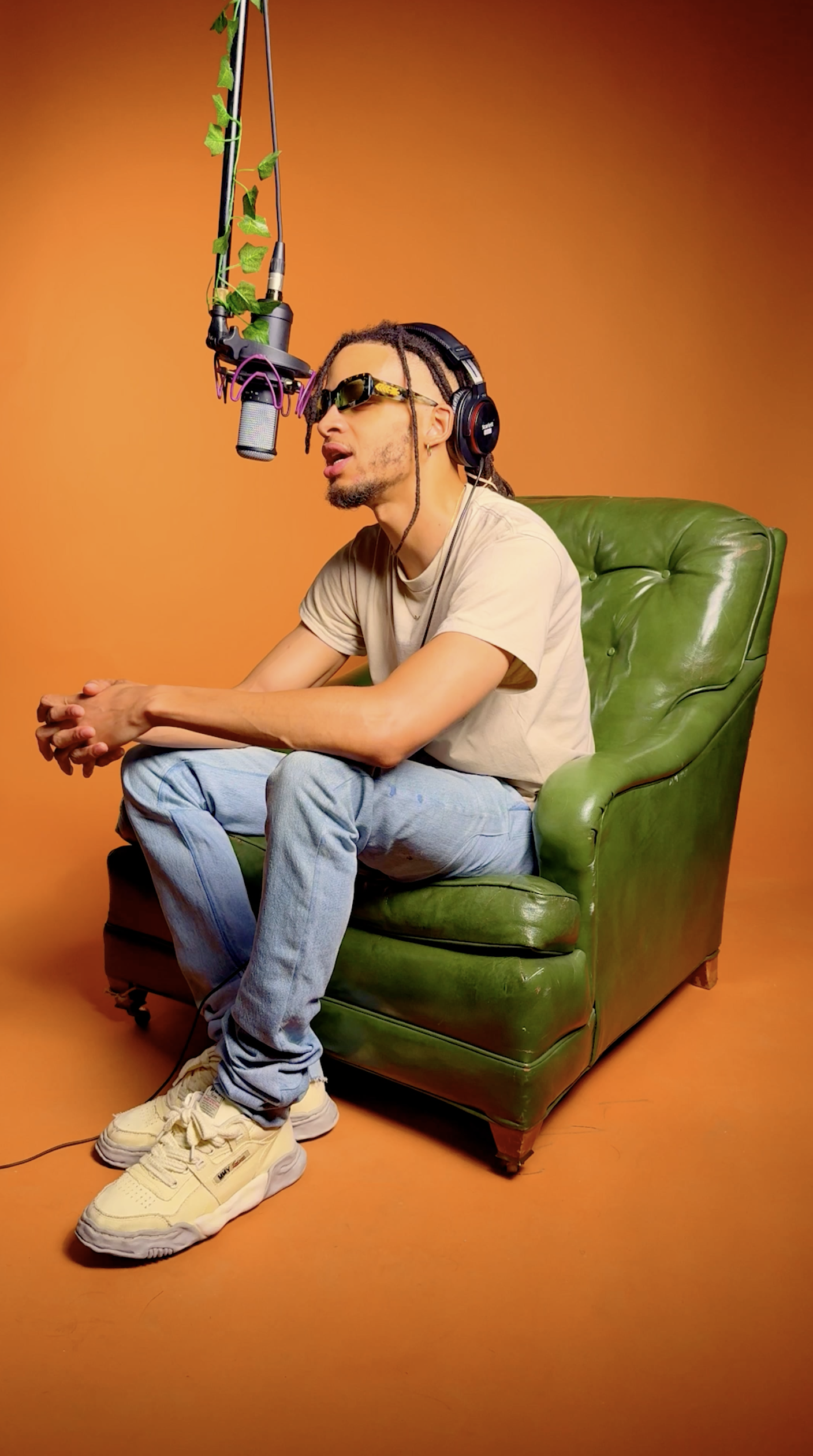 A man with braids and sunglasses sitting on a green leather armchair in front of an orange background, singing into a microphone suspended from a boom stand.