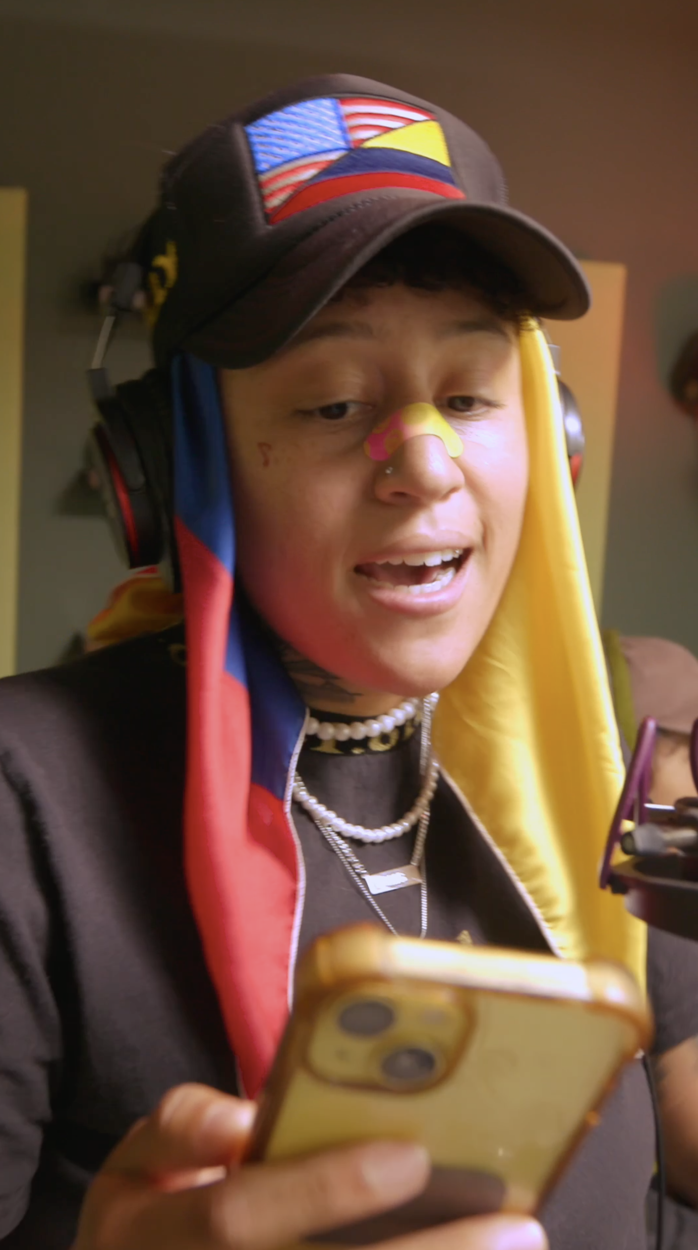 A person wearing a black cap with an American flag patch, headphones, a nose bandage, and layered necklaces, looking at a smartphone.