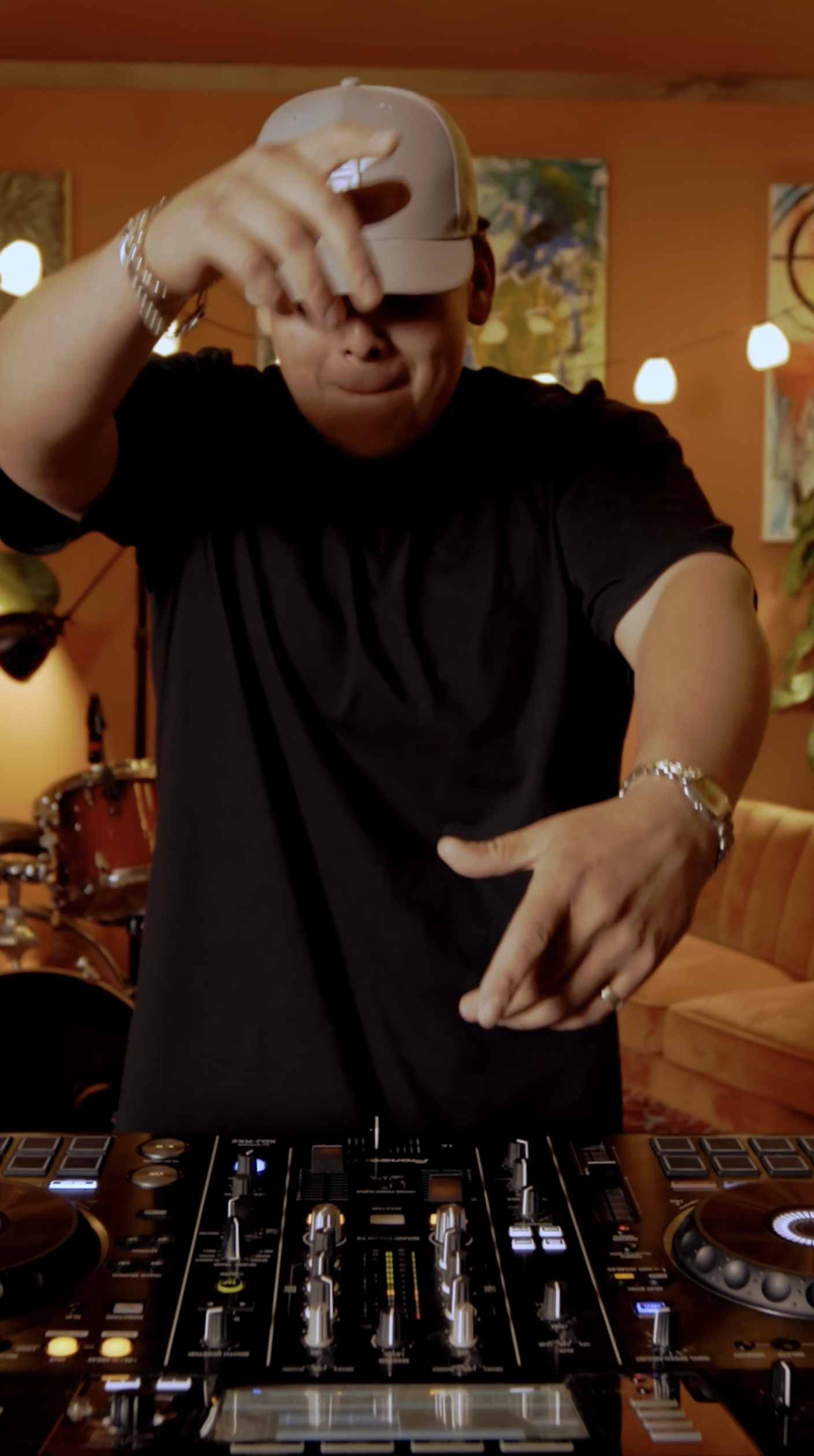 A musician wearing a black t-shirt, white cap, and silver jewelry, DJing with headphones and a DJ console in a warmly lit indoor setting with colorful paintings and string lights.