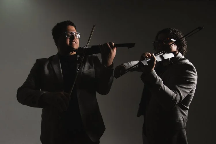 Two men in suits playing violins in silhouette against a gray background