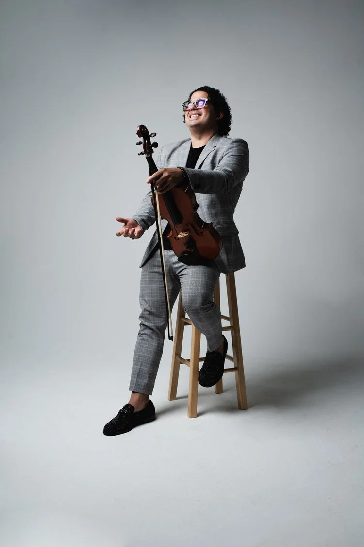 Man in gray plaid suit sitting on a stool, holding a violin and smiling, against a plain gray background.