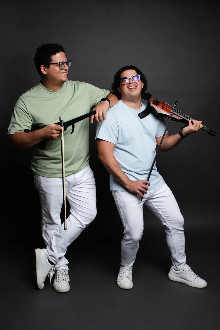 Two young men are posing with musical instruments, one with a violin and the other with a bow, against a black background. They are smiling and wearing casual light-colored clothing and glasses.