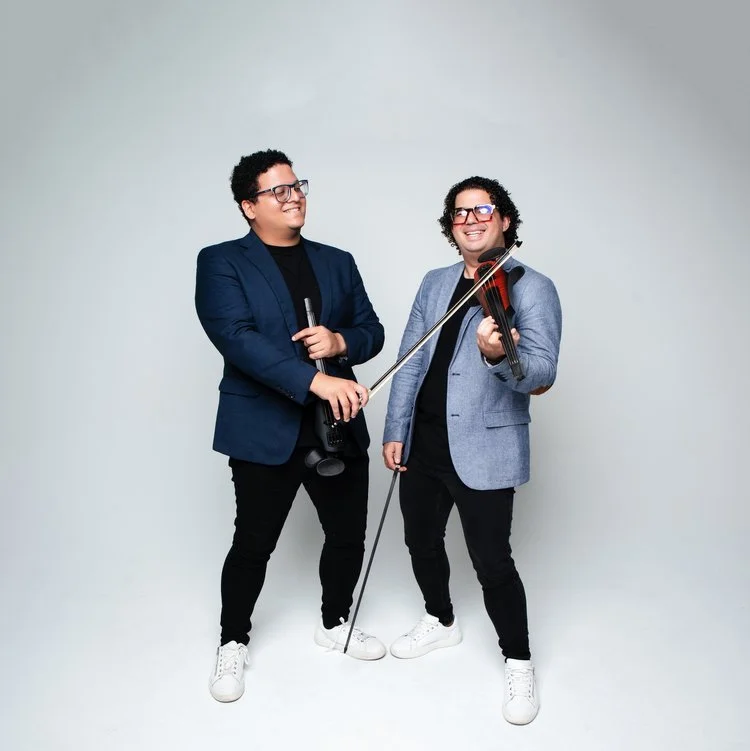 Two men in casual business attire, one holding a microphone and the other playing a violin, standing and smiling against a plain gray background.
