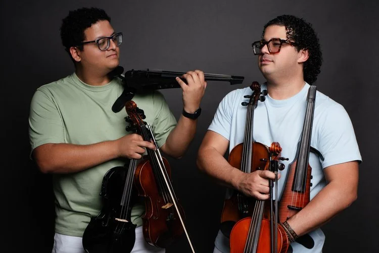 Two young men holding violins and a viola against a dark background, one man is playing a virtual violin held to the other man's face.