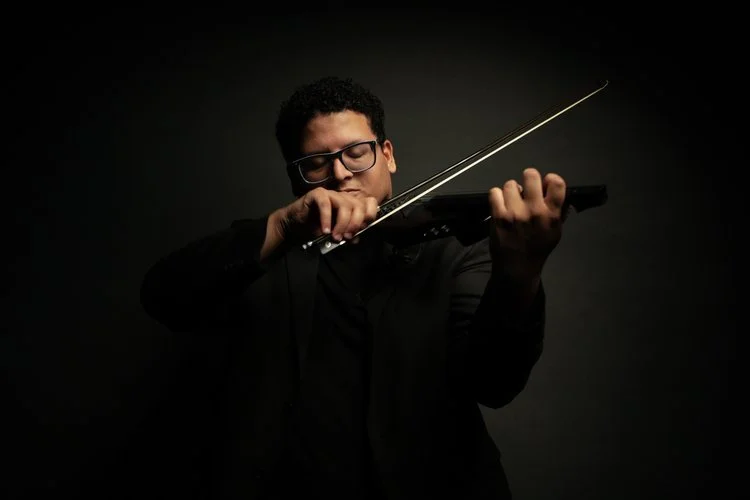 A person wearing glasses playing an electric violin in a dark setting.
