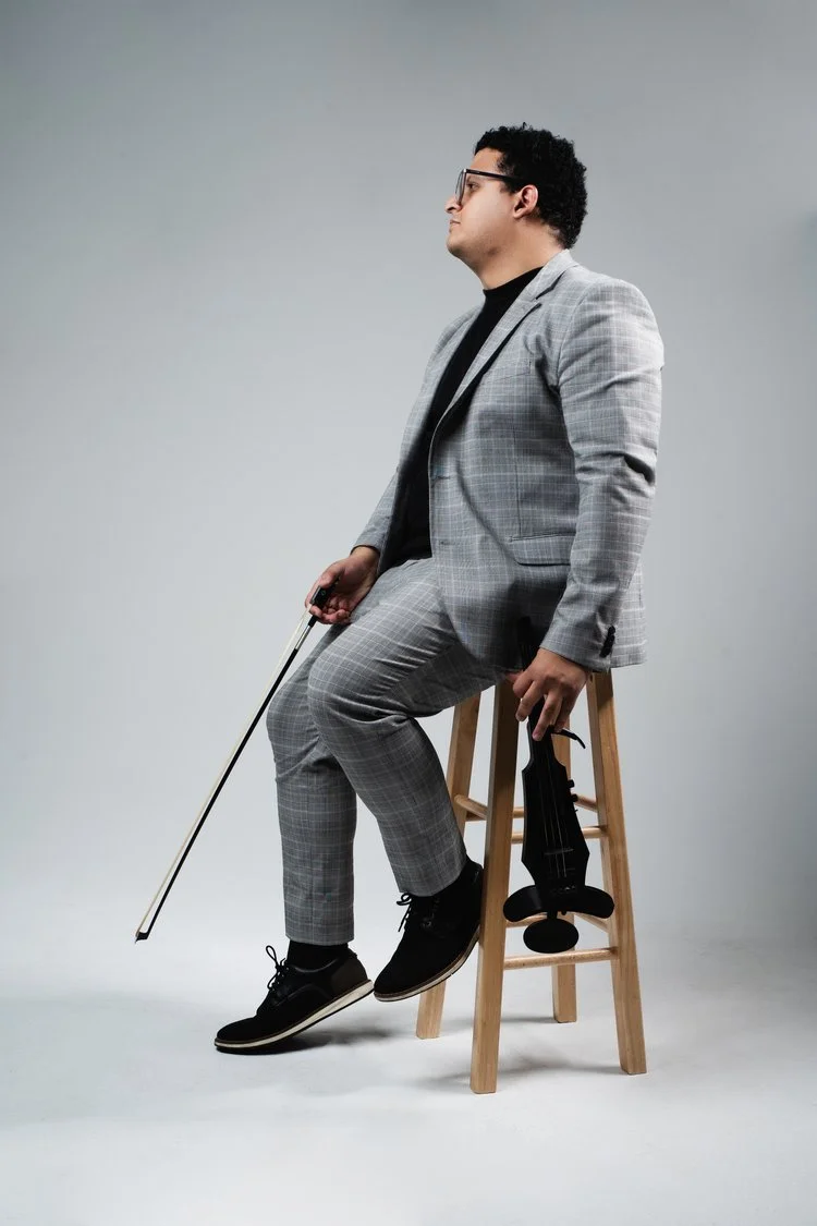 Young man in a gray plaid suit sitting on a wooden stool, holding a violin and bow, with a gray background.